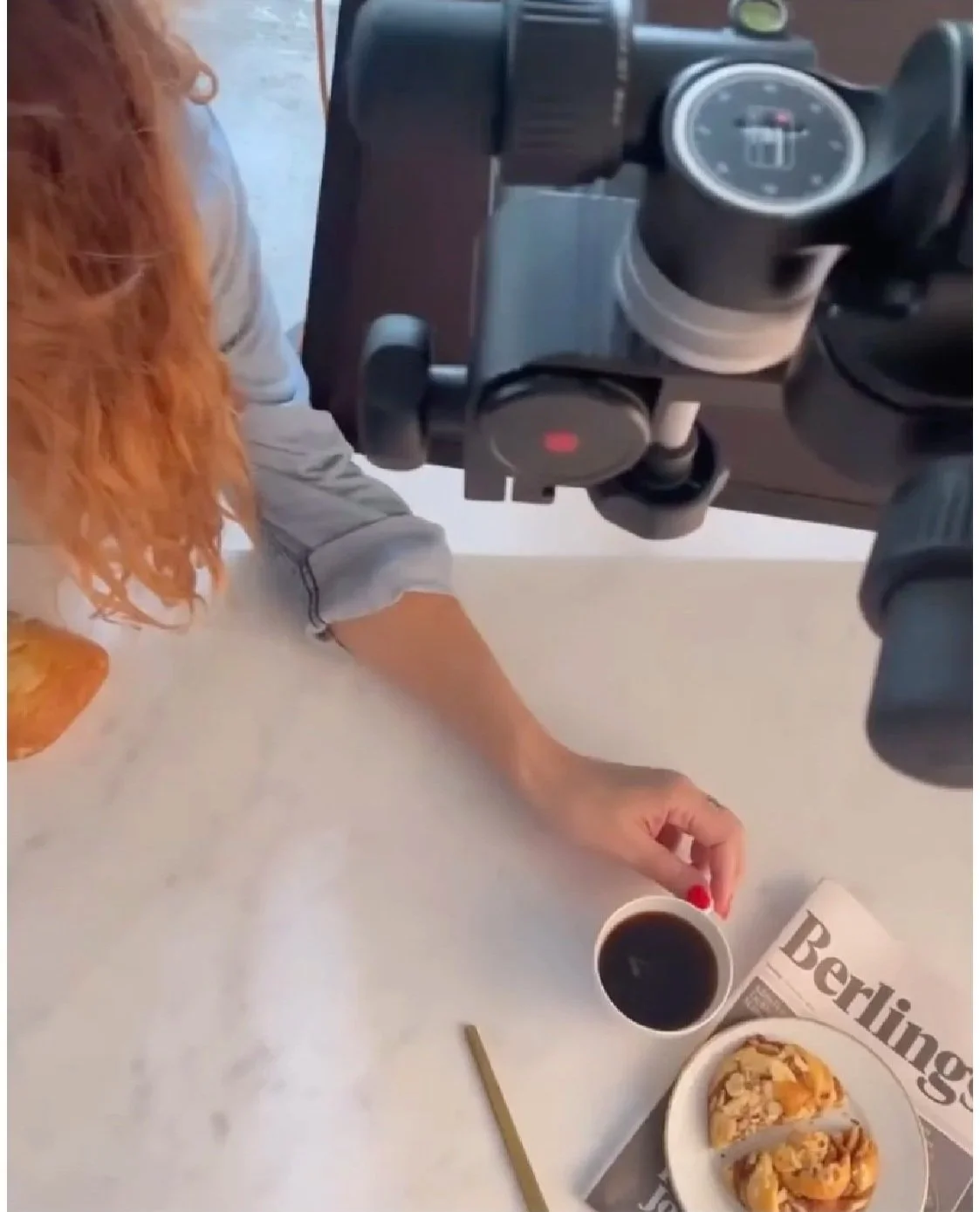 En person med rødt hår, tager et billede af en kop kaffe og en smule croissanter på en tallerken, set fra oven, med en fotografisk kamerahøjde.