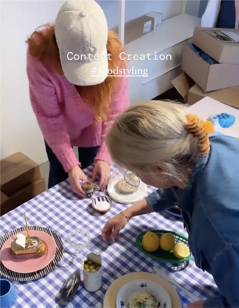 To kvinder står ved et bord dækket med en blå og hvid ternet duk. Der er kage, citroner, slik og kopper på bordet. Kvinden til venstre har rødbrunt hår, en pink sweater og en bandage med hætte på hovedet. Kvinden til højre har blond hår og er iført denim.