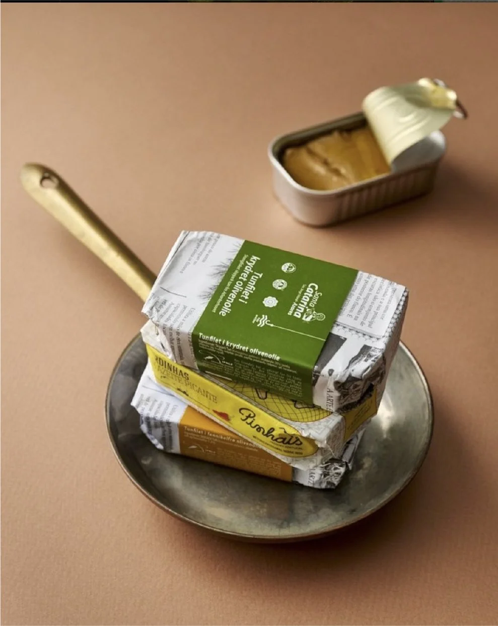 To pakker med fisk, en åben dåse med sild, en kniv og en metalbakke på et bord.