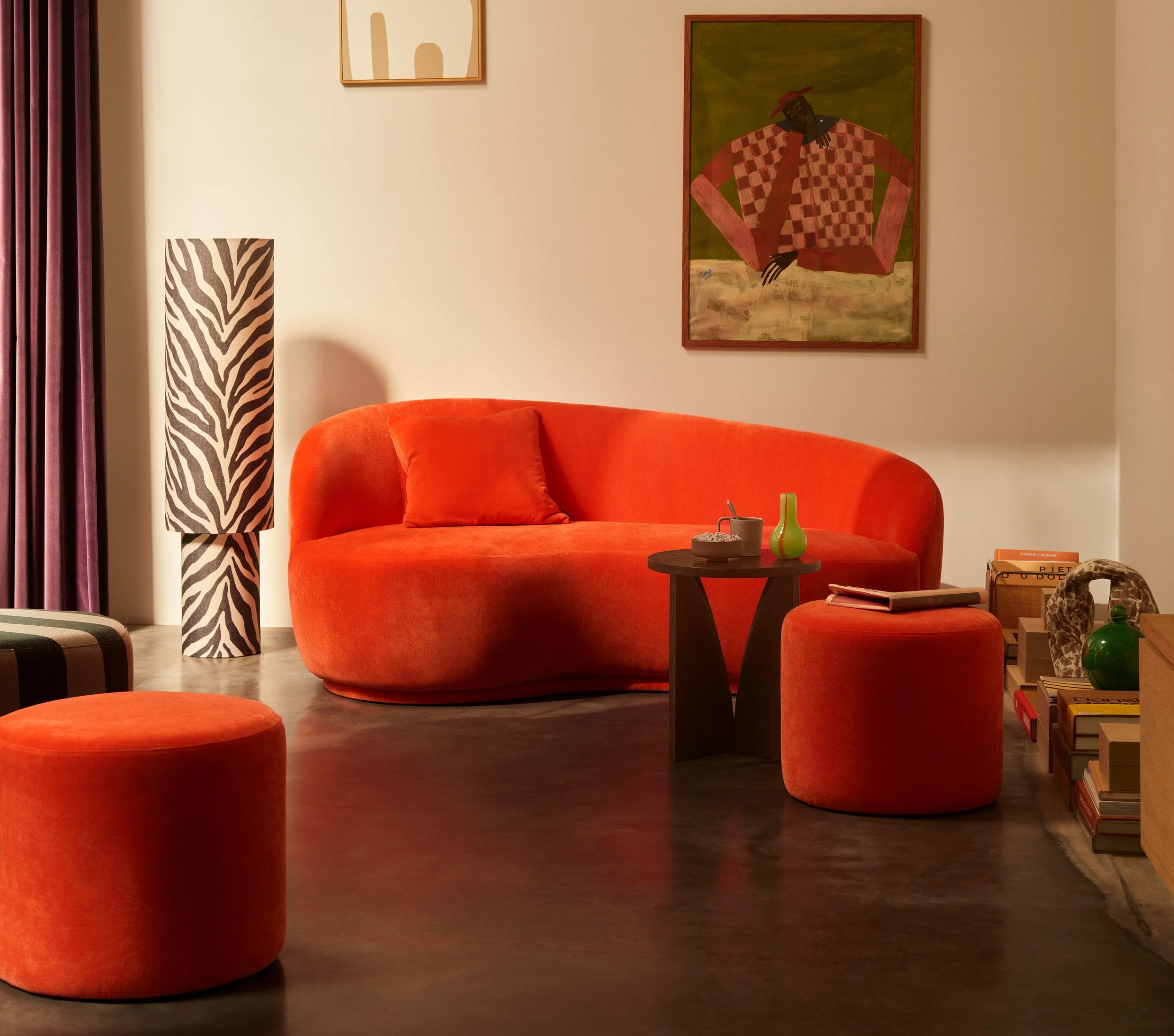 En moderne stue med et orange sofa, matchende puf, et rundt sort bord med små dekorationer, en zebra-print gulvlampe, kunst på væggene, og en række bøger og skulpturer på et lavt møbel.