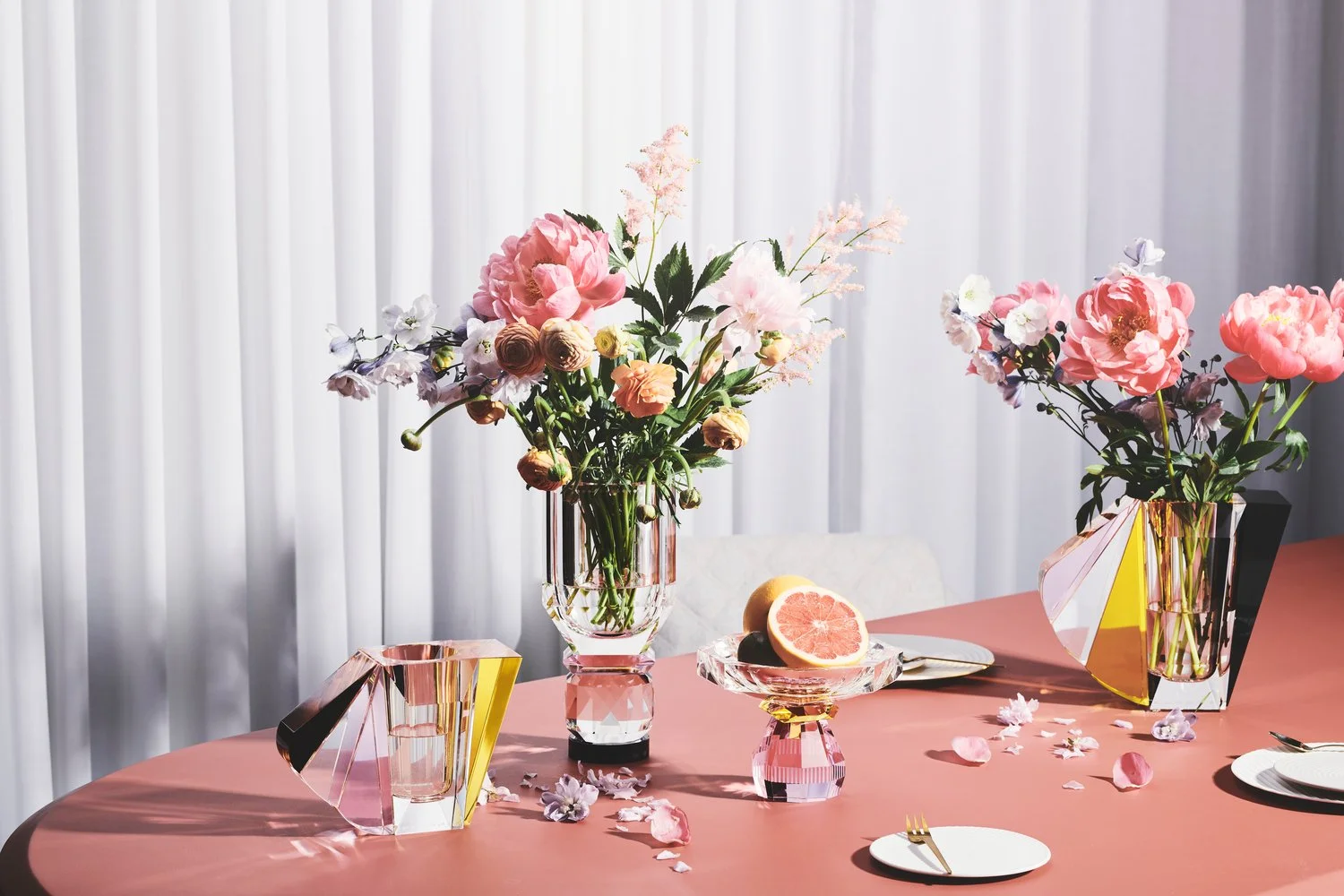 Bord med lyserøde blomsterarrangementer i gennemsigtige vaser, en skål med et skåret grapefruit, urtepinde, tallerkener med bestik, og et par frugt, på en pink borddække.