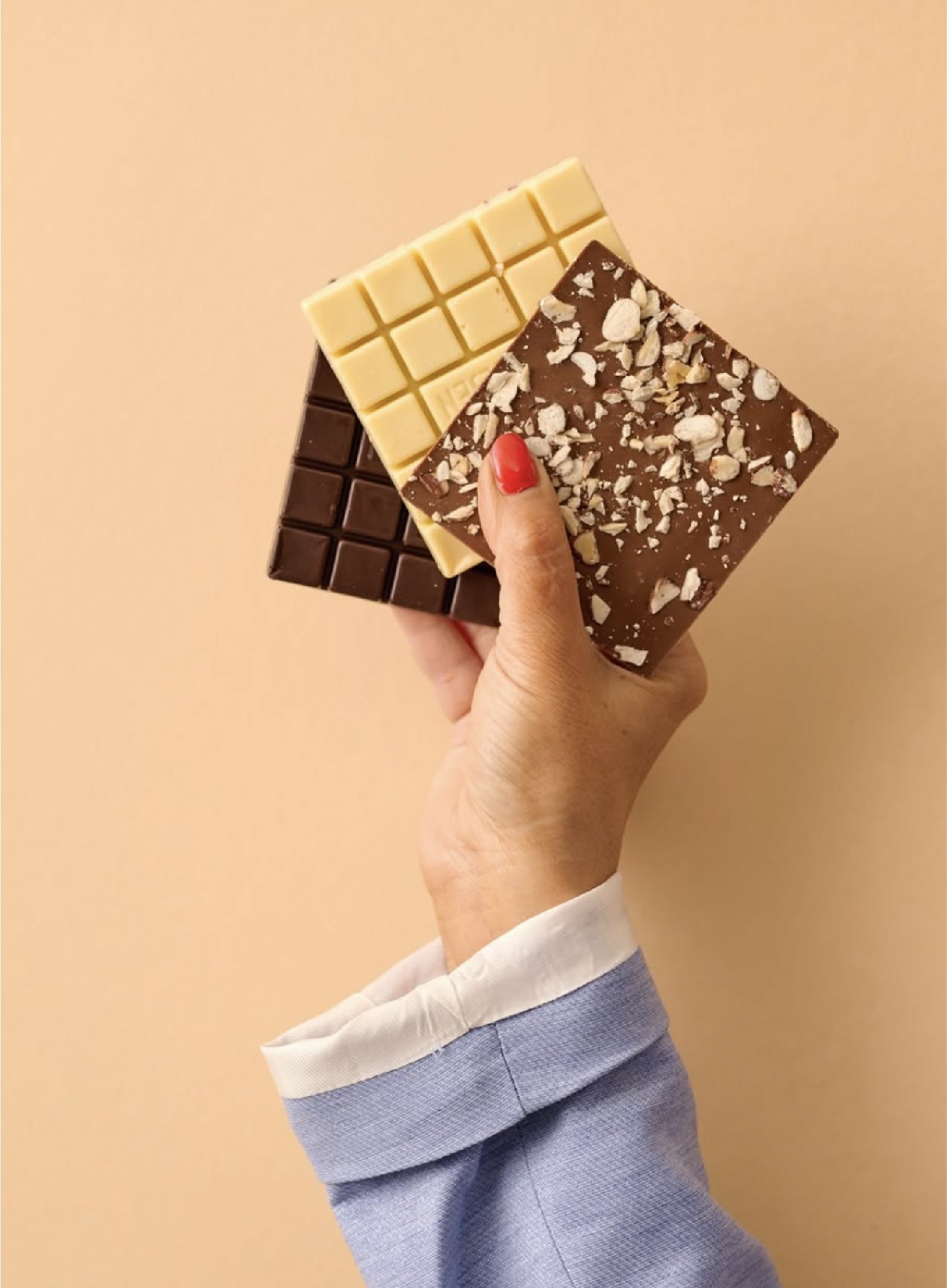 En hånd holder tre stykker chokolade med forskellige smagsvarianter og toppings, mod en beige baggrund.