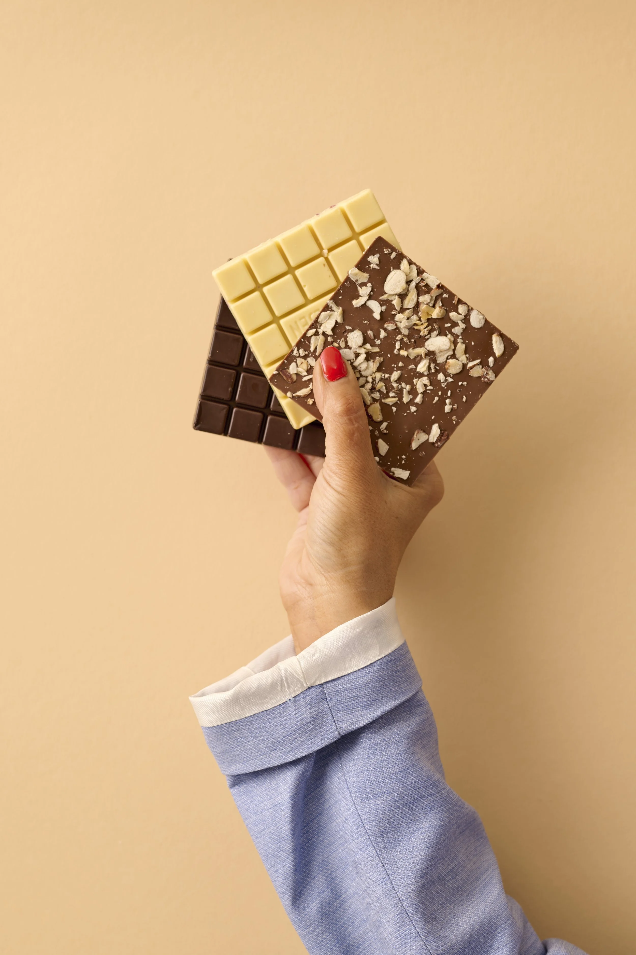 Hånd med rød neglelak holder tre forskellige chokoladestykker mod en lysebrun væg. Chokoladen er hvid, mælke og mørk chokolade, hvor den med nødder er toppet.