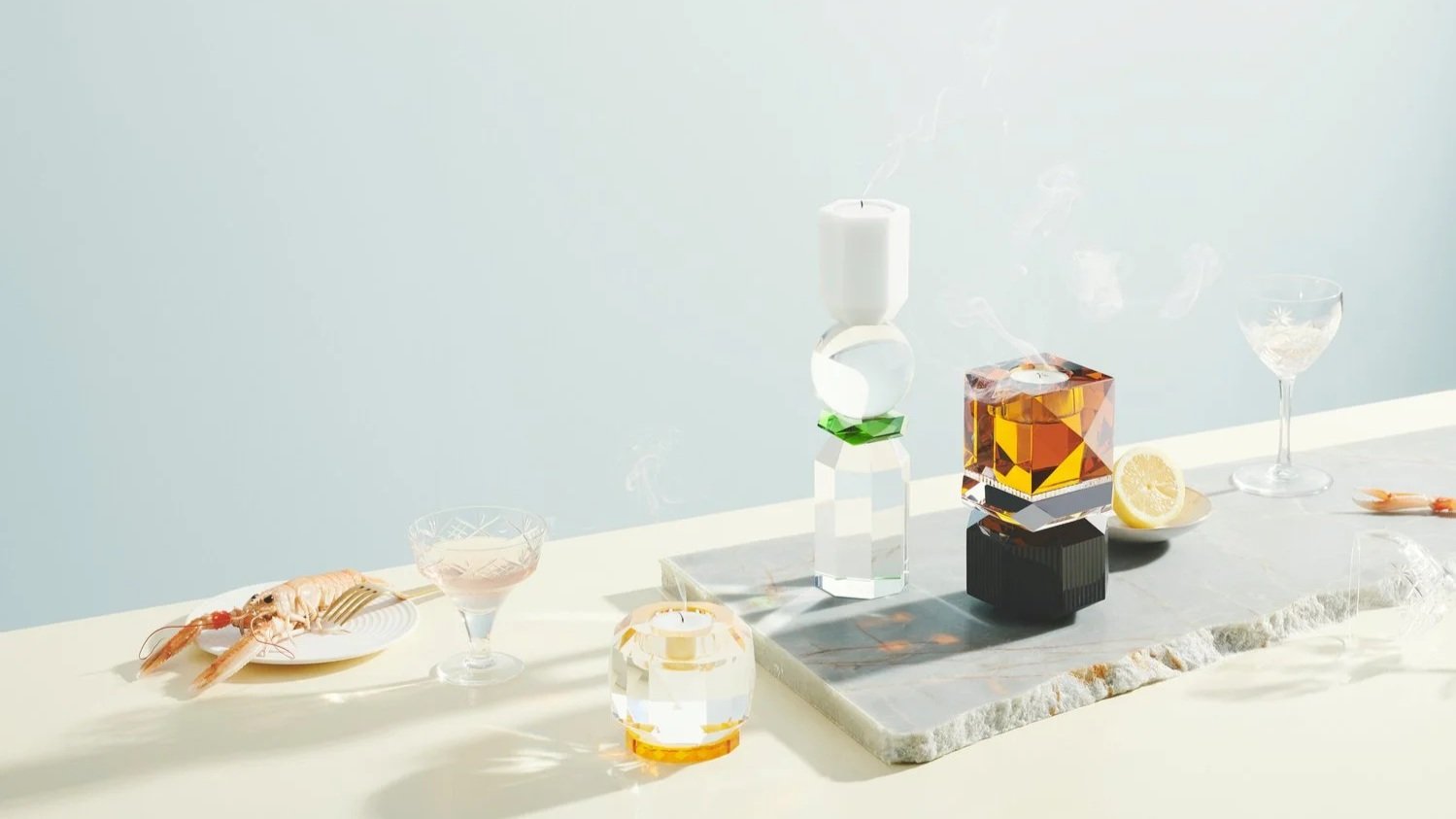 En minimalistisk borddækning med glas, stearinlys, citron og en hvid tallerken med en reje, sat op på en lys overflade.