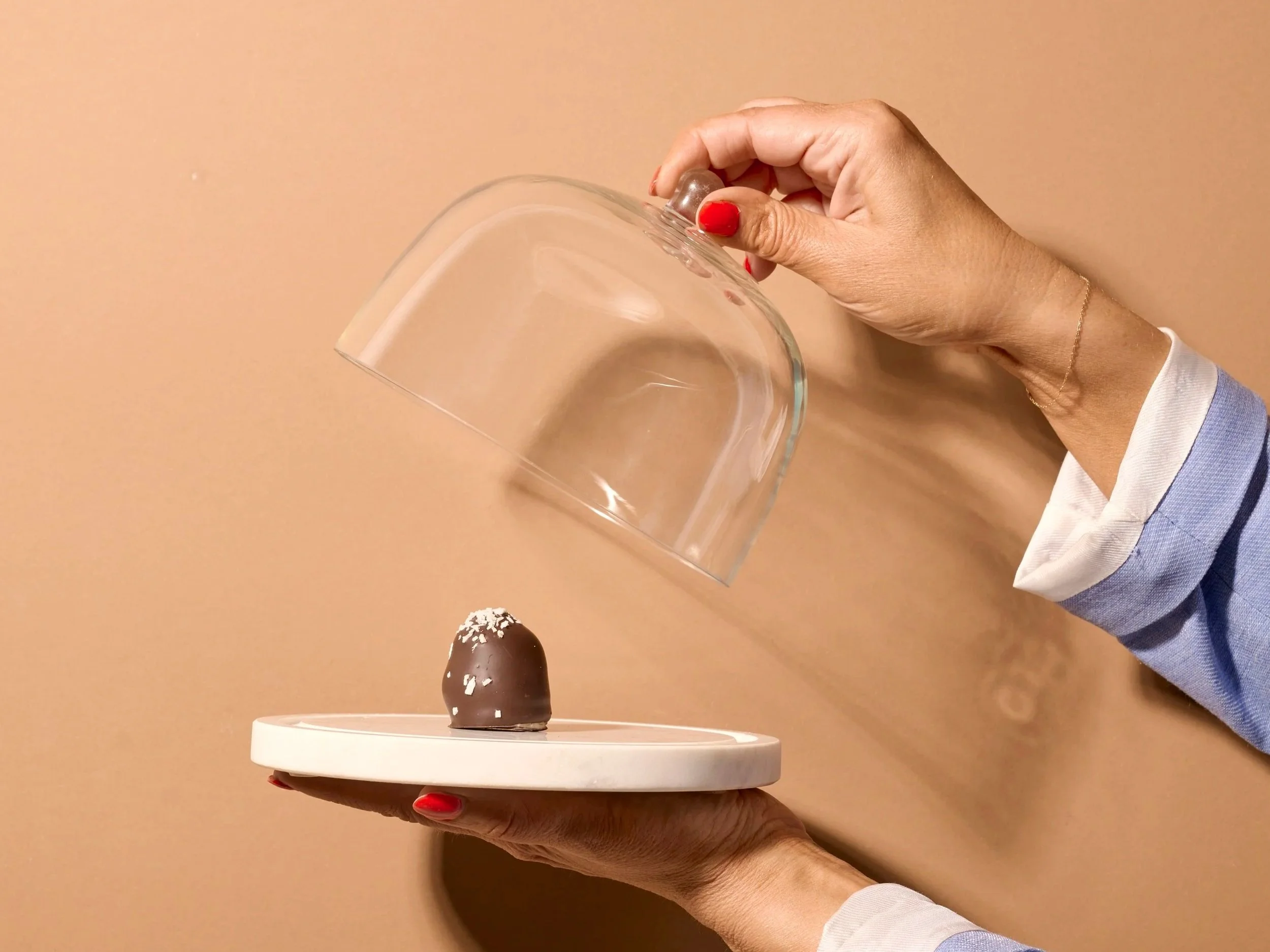 Hænder holder en afrundet chokolade på en hvid tallerken, mens den bliver dækket med en gennemsigtig skål.