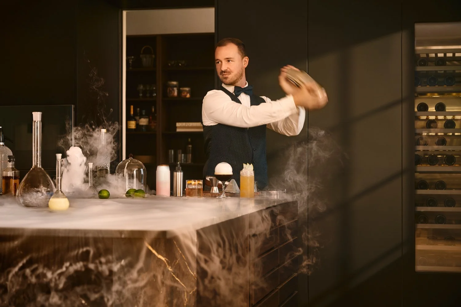 En bartender, der shaker en cocktail i et moderne, mørkt køkken med stilfulde opbevaringsskabe og laboratorie-agtige glaseringer, omgivet af dry ice og ingredienser på bordet.