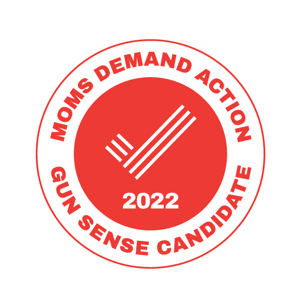 Red circular badge with black text and a black symbol of three parallel lines and three shorter lines intersecting diagonally, with the year 2022 at the bottom. The text reads 'MOI'S DEMAND ACTION GUN SENSE CANDIDATE' around the circle.