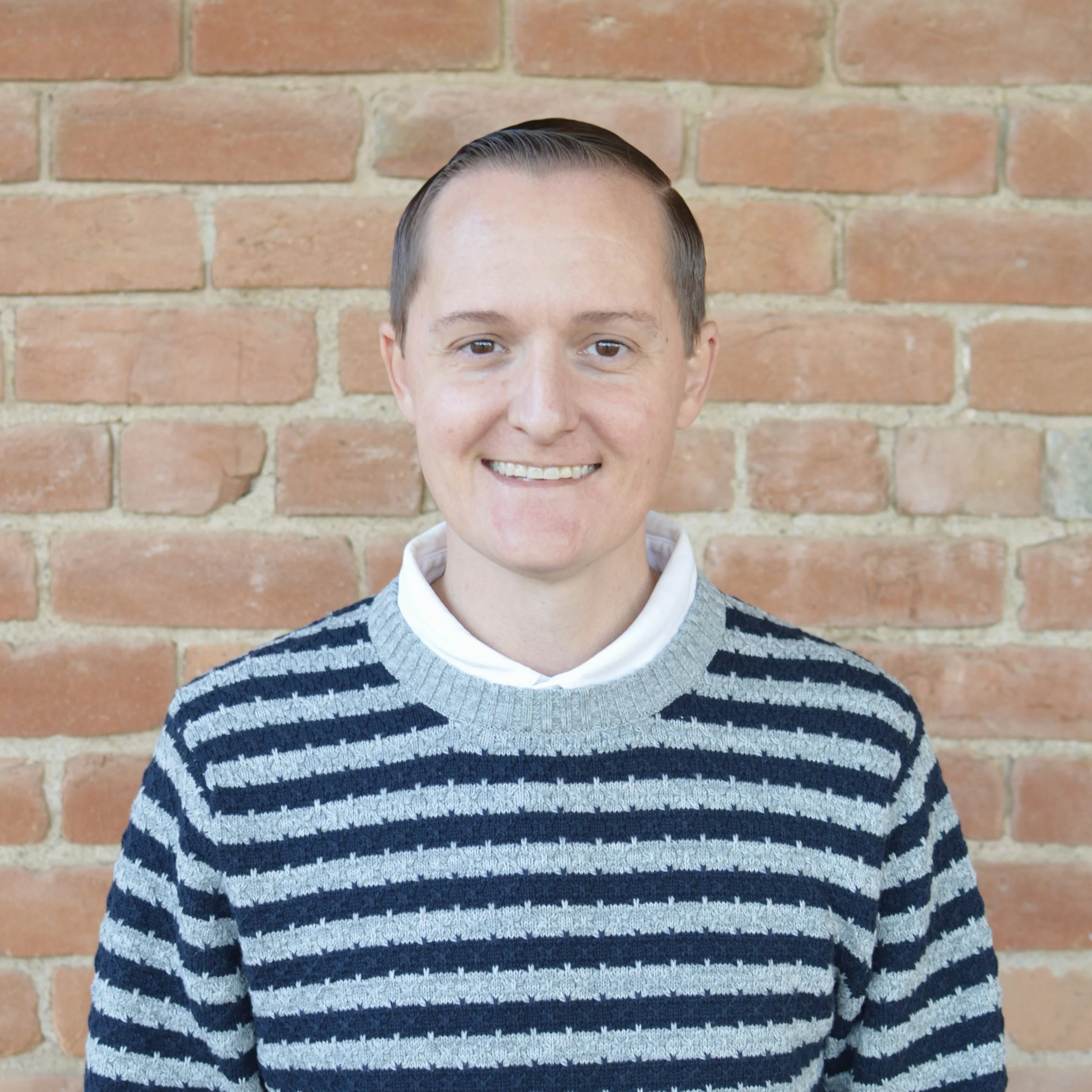 Candidate Kyle Clayton standing in front of a brick wall with a black and gray striped sweater