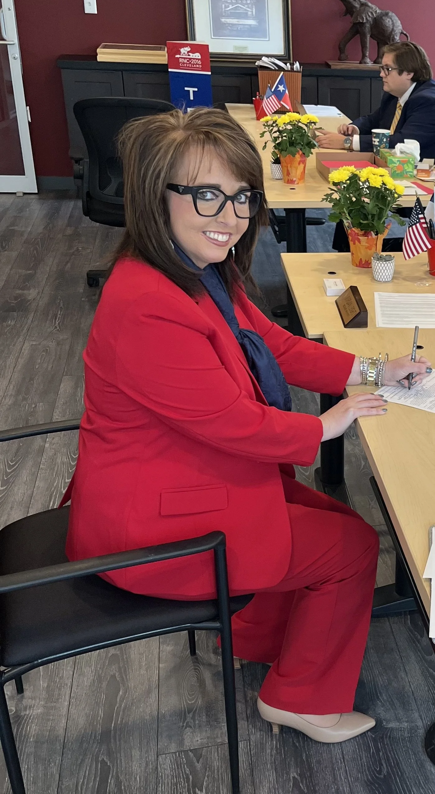 Rachel Hogue with brown hair, glasses, wearing a red suit and beige heels, is sitting at a conference table, smiling and filing her candidacy.