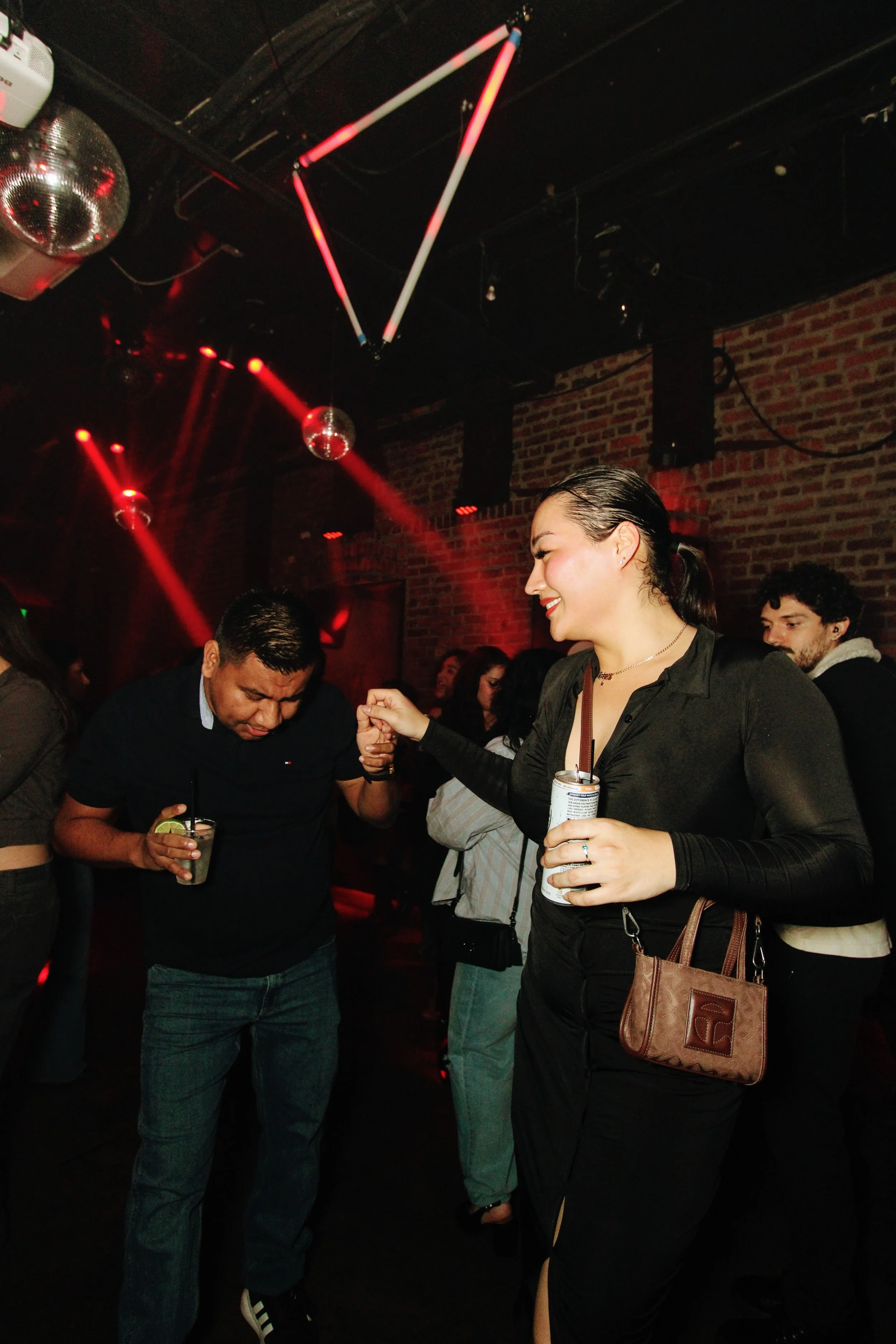 People dancing and socializing in a nightclub with red lighting, disco balls, and a brick wall background.