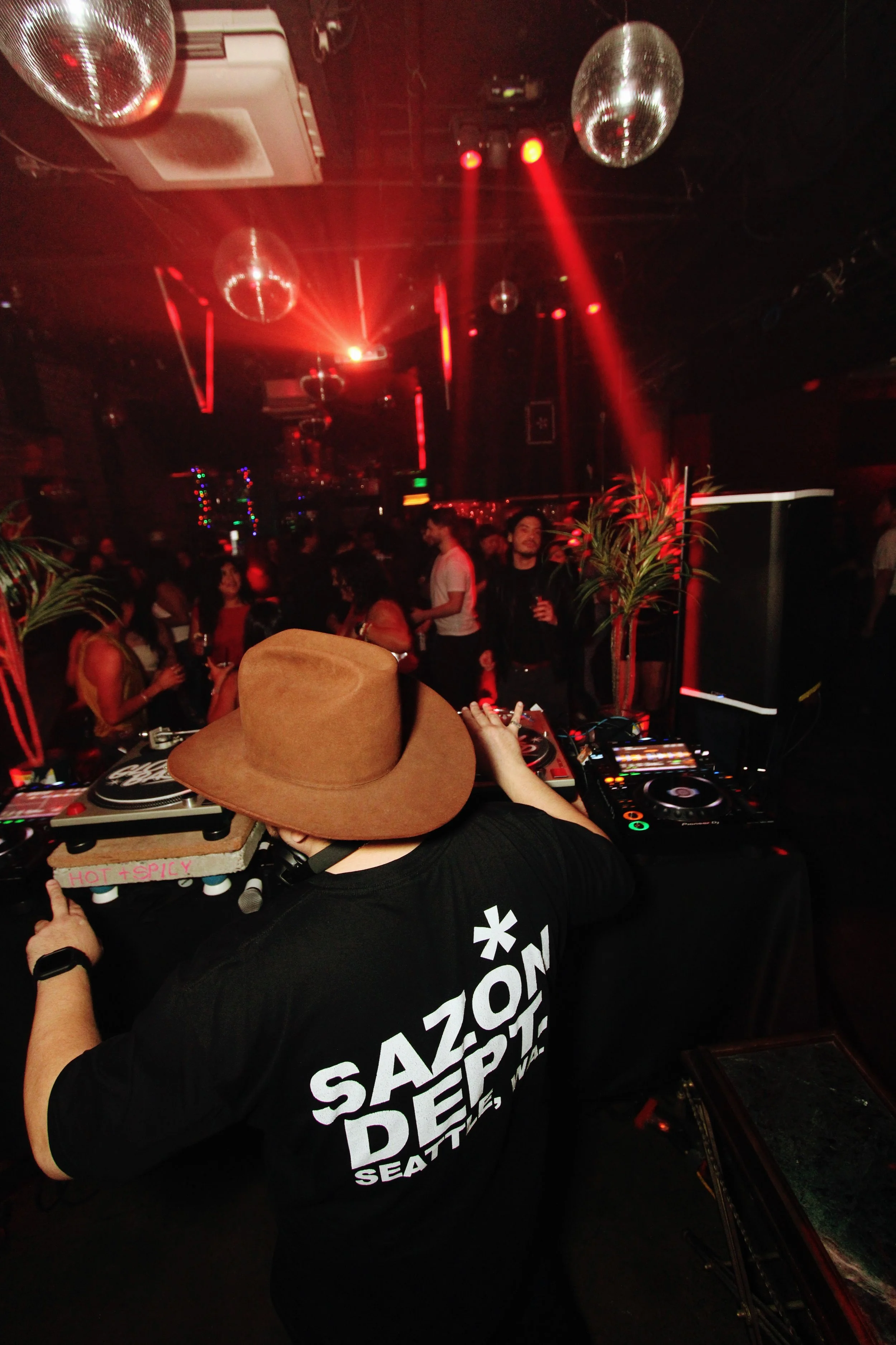 DJ wearing a brown hat and a black t-shirt with white text, performing at a nightclub with a crowd dancing under red lighting and disco balls.