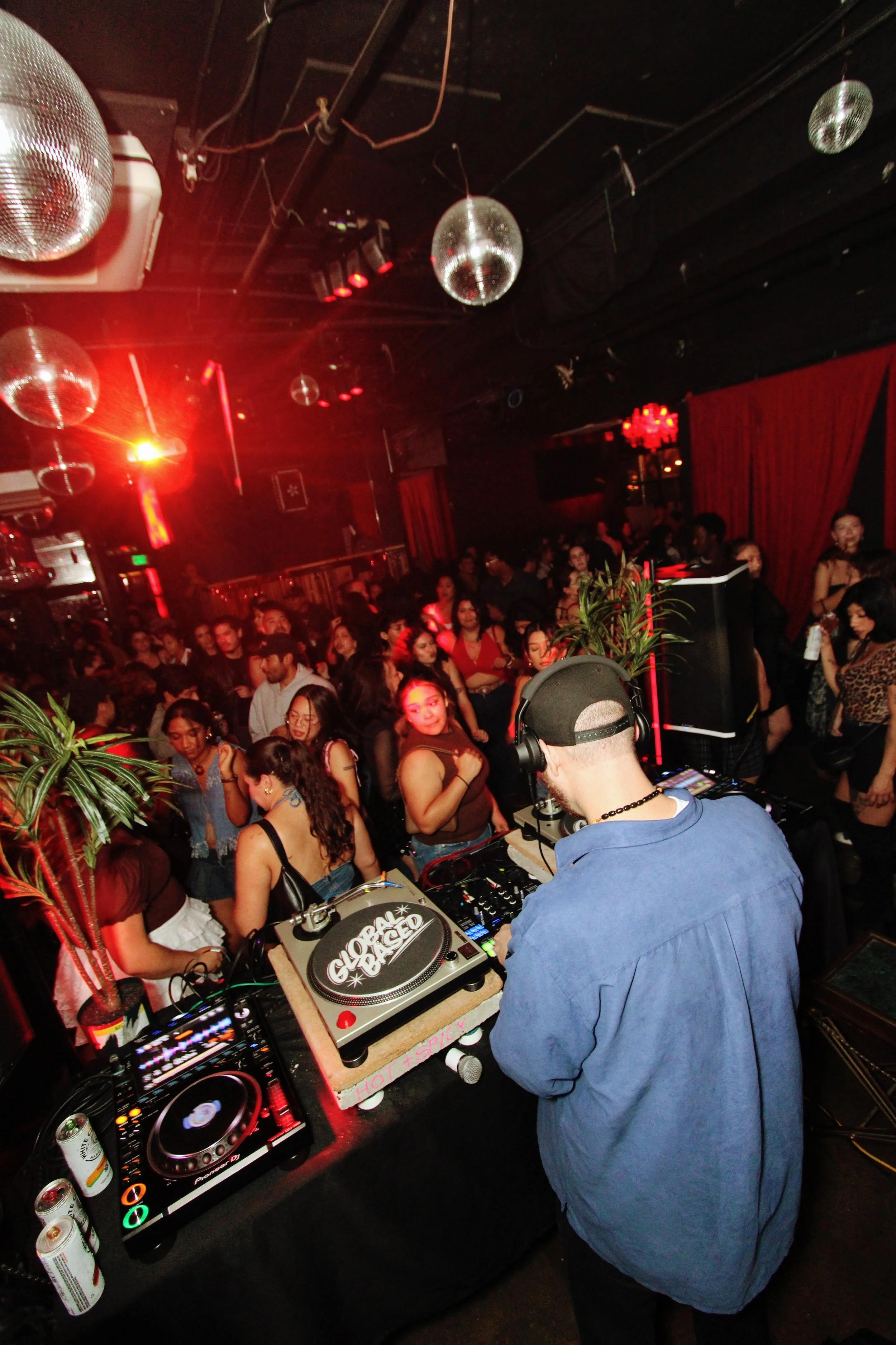 A DJ is performing at a nightclub with a large crowd dancing. Disco balls hang from the ceiling, and red lighting illuminates the scene.
