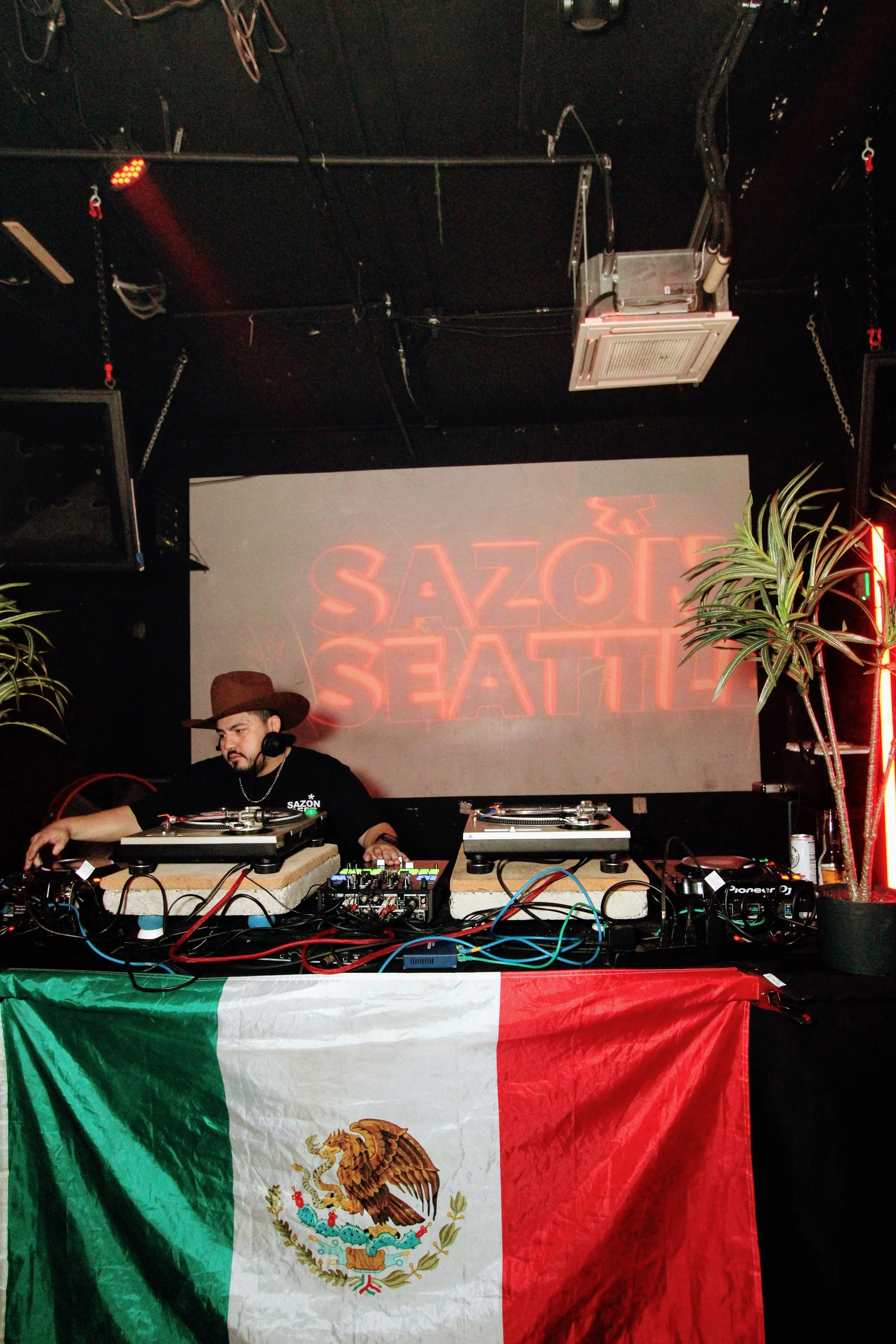 A DJ wearing a brown hat and black t-shirt playing music behind a booth decorated with a Mexican flag. The background features a screen with red neon text that says 'Sazon Seattle' and two potted plants.