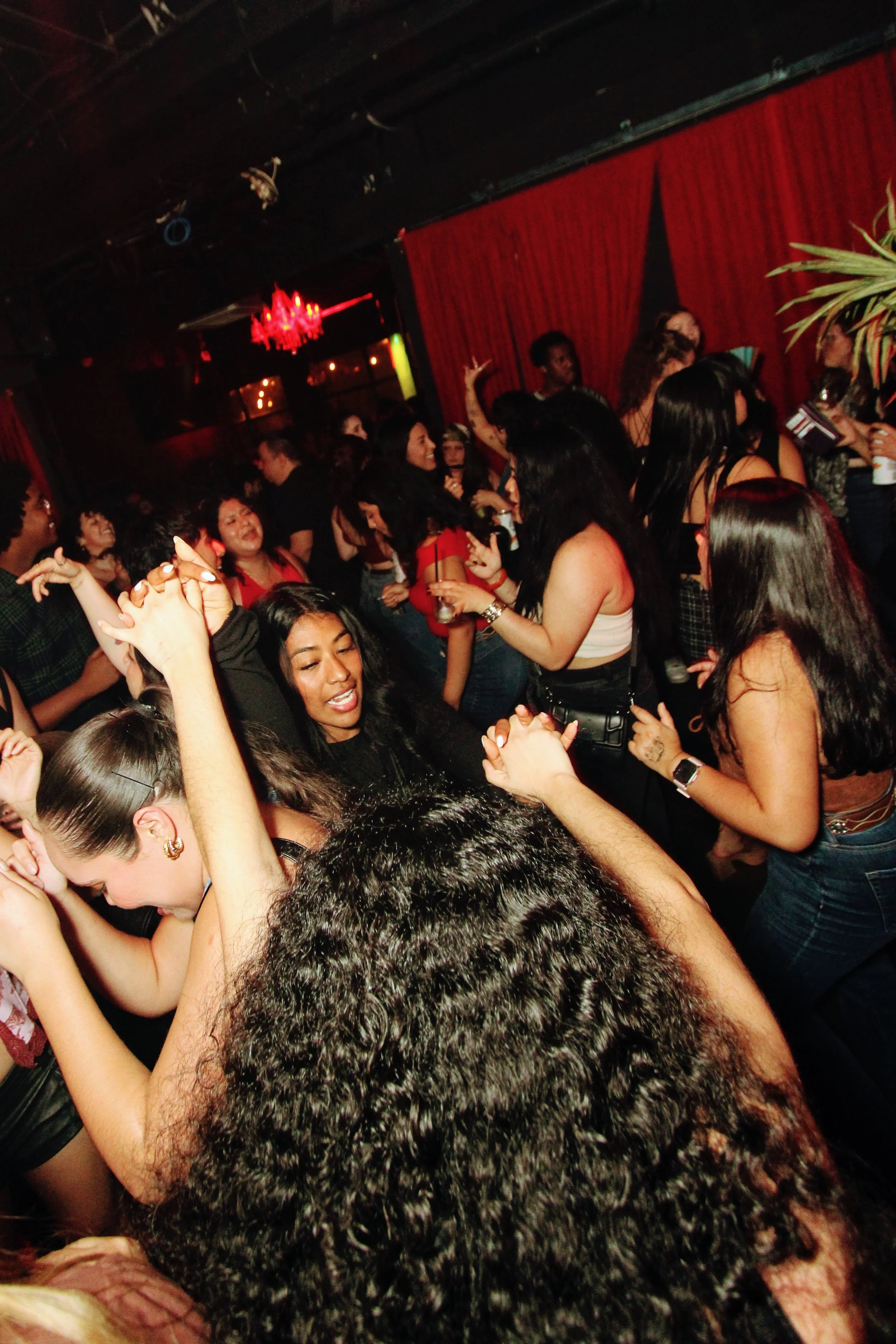 People dancing and socializing inside a club with red curtains and dim lighting.