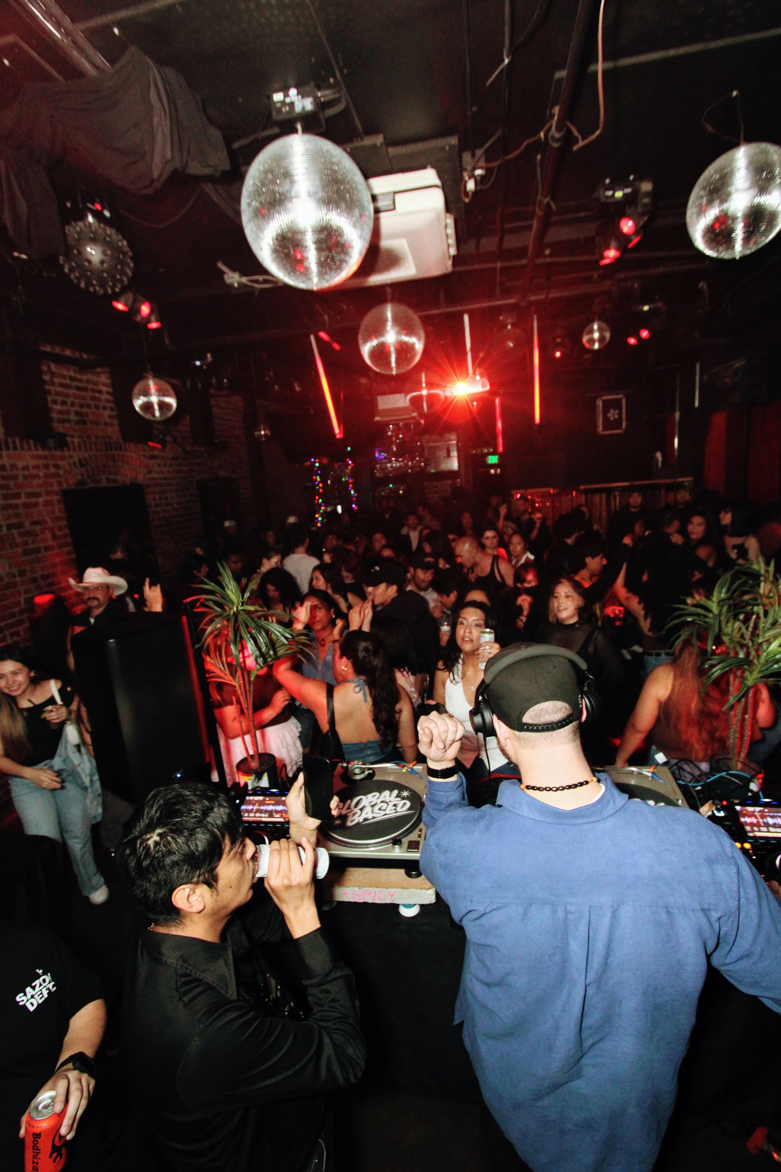 Crowd of people dancing and socializing in a nightclub with disco balls and red lighting, a DJ mixing music at the turntables.