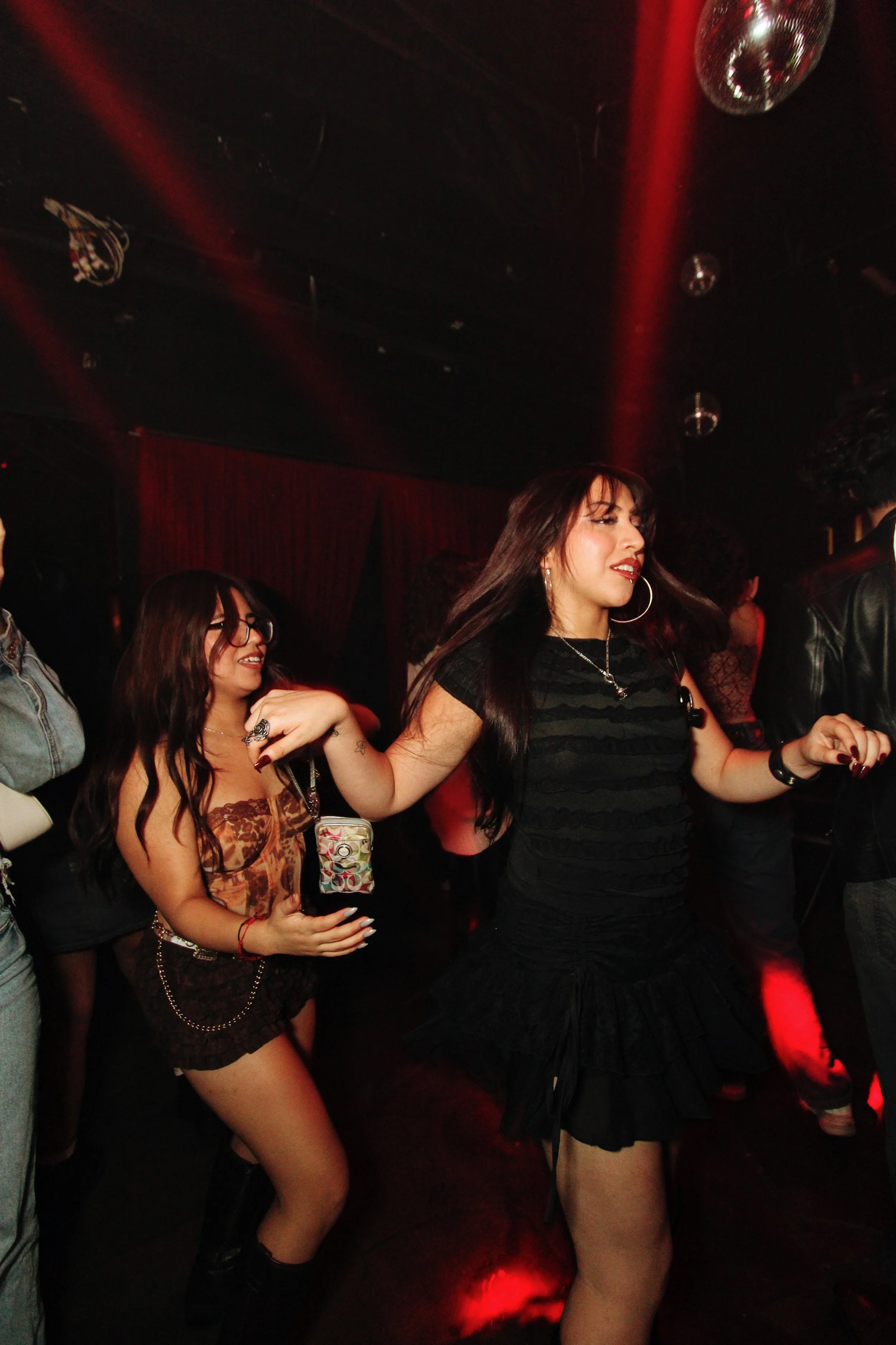 Two women dancing in a nightclub, one in a black dress with hoop earrings and a necklace, the other in a strapless top and shorts, both smiling and holding hands.