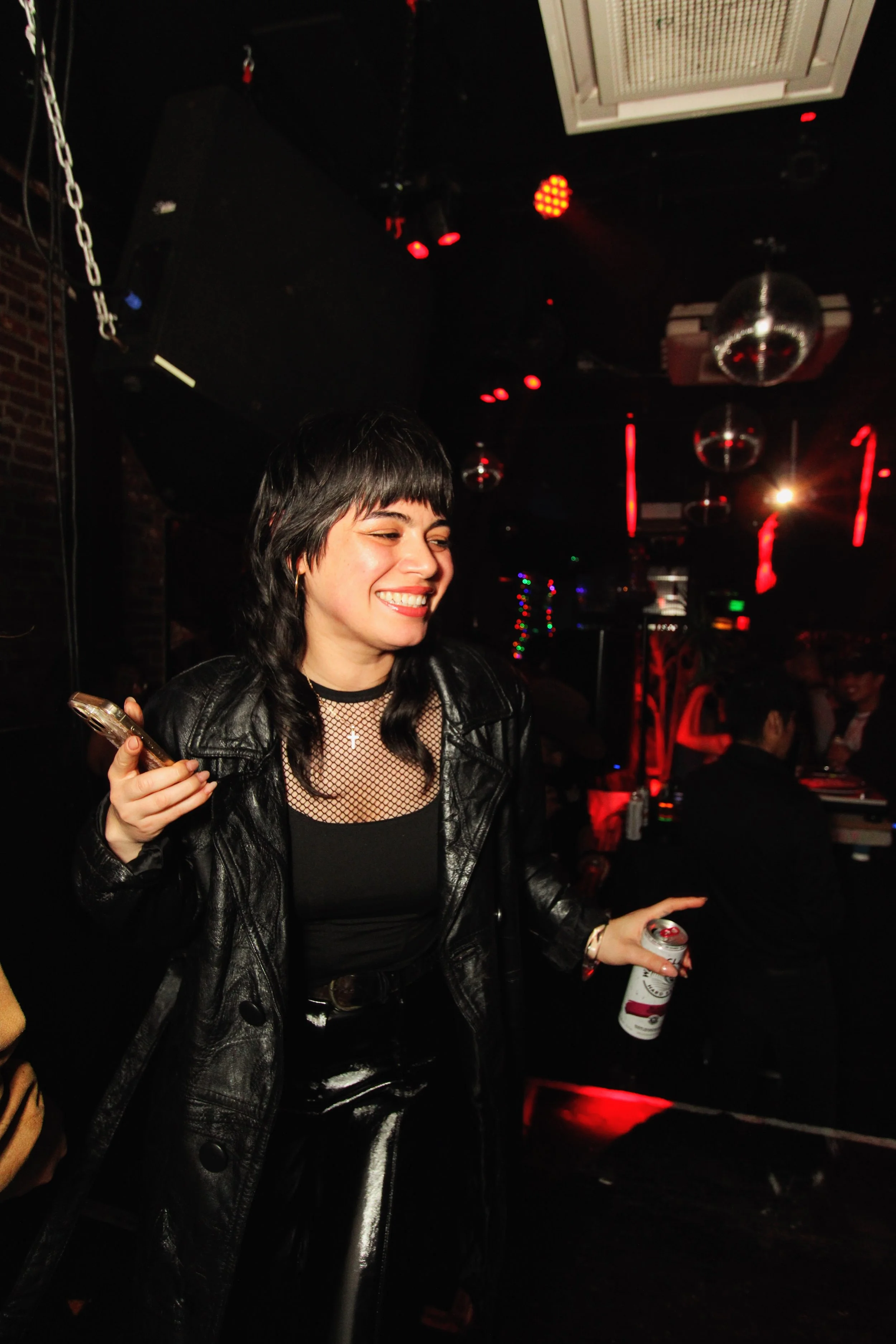 A woman dancing and smiling at a nightclub, holding a cellphone in one hand and an open can of beer in the other, with red and black lighting and disco balls overhead.
