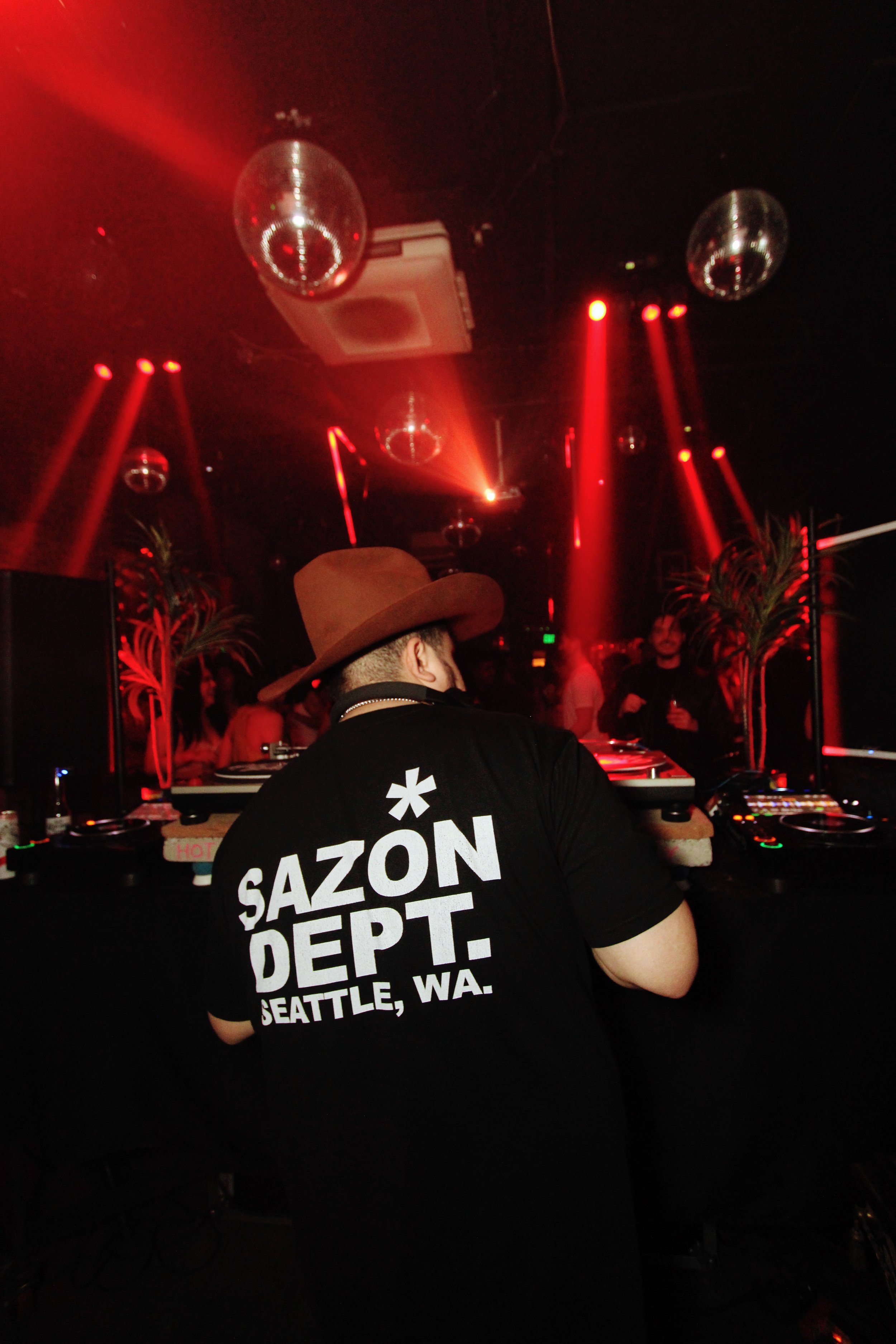 A DJ wearing a brown hat and a black T-shirt with white lettering, playing music at a club with red lighting and disco balls hanging from the ceiling, and a few people in the background.