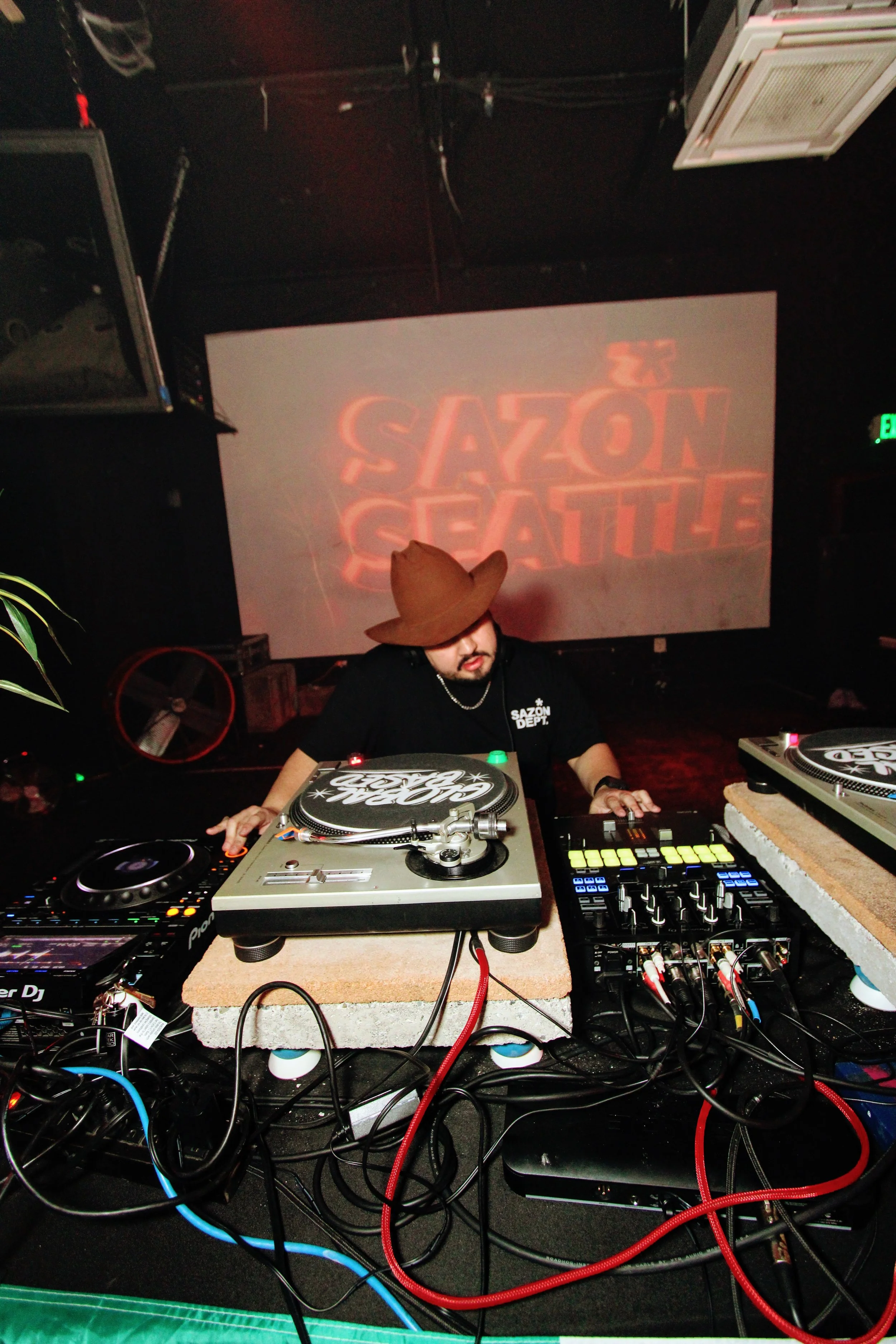 A DJ wearing a cowboy hat is performing in a dimly lit club. There are turntables, mixers, and wires in front of him. A large screen behind displays the text 'SAZON SEATTLE' in red neon style.