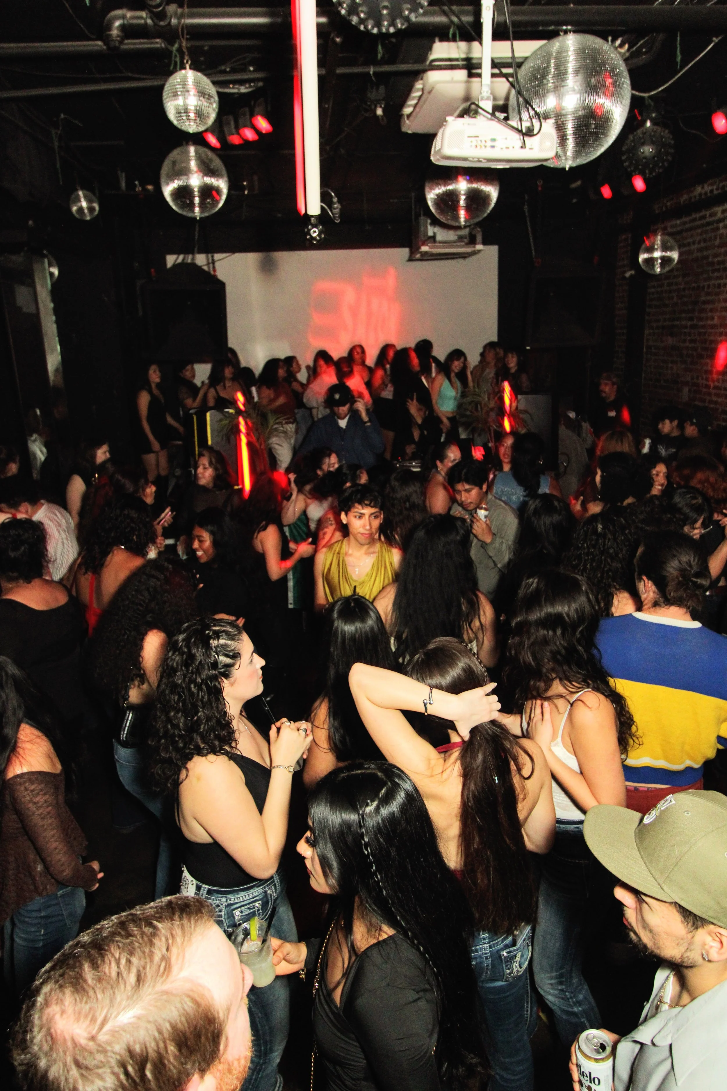 Crowded dance floor with people dancing at a nightclub, disco balls hanging from the ceiling, and colorful lighting.