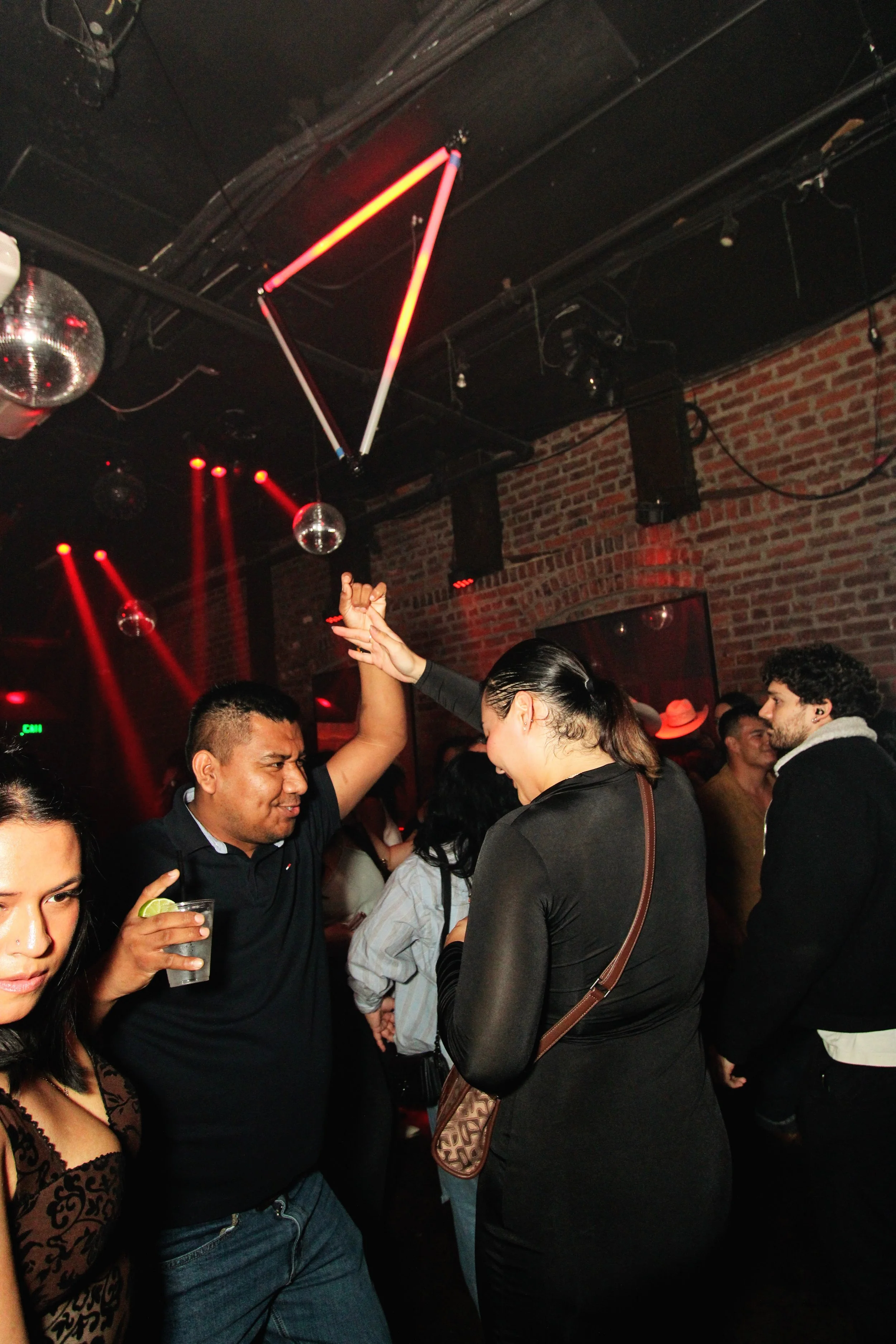 People dancing and socializing in a dimly lit nightclub with red lighting, brick walls, disco balls, and a geometric neon ceiling light.