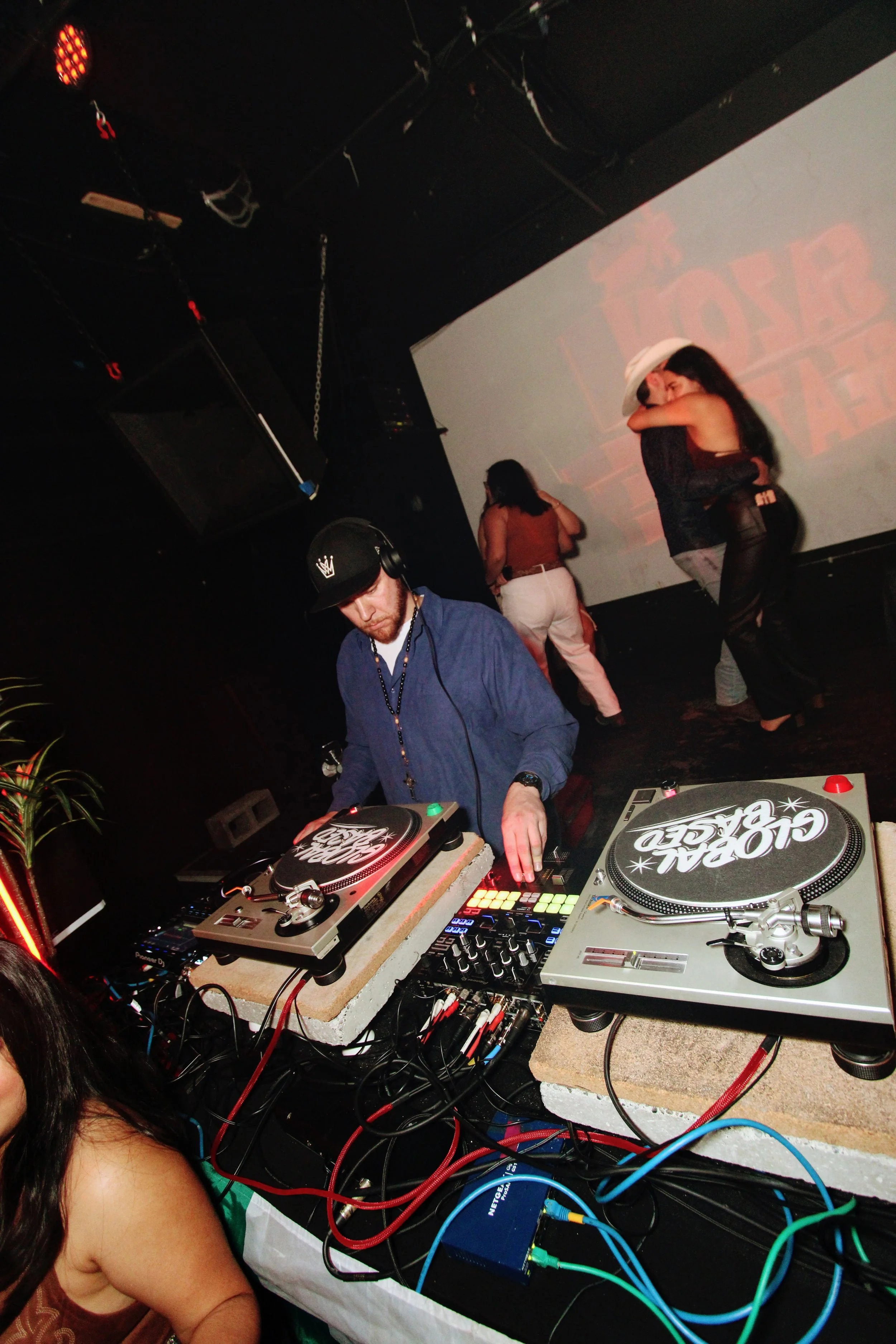 A DJ wearing headphones and a baseball cap playing music on turntables with a mixer at a party, with two women dancing on a stage in the background.