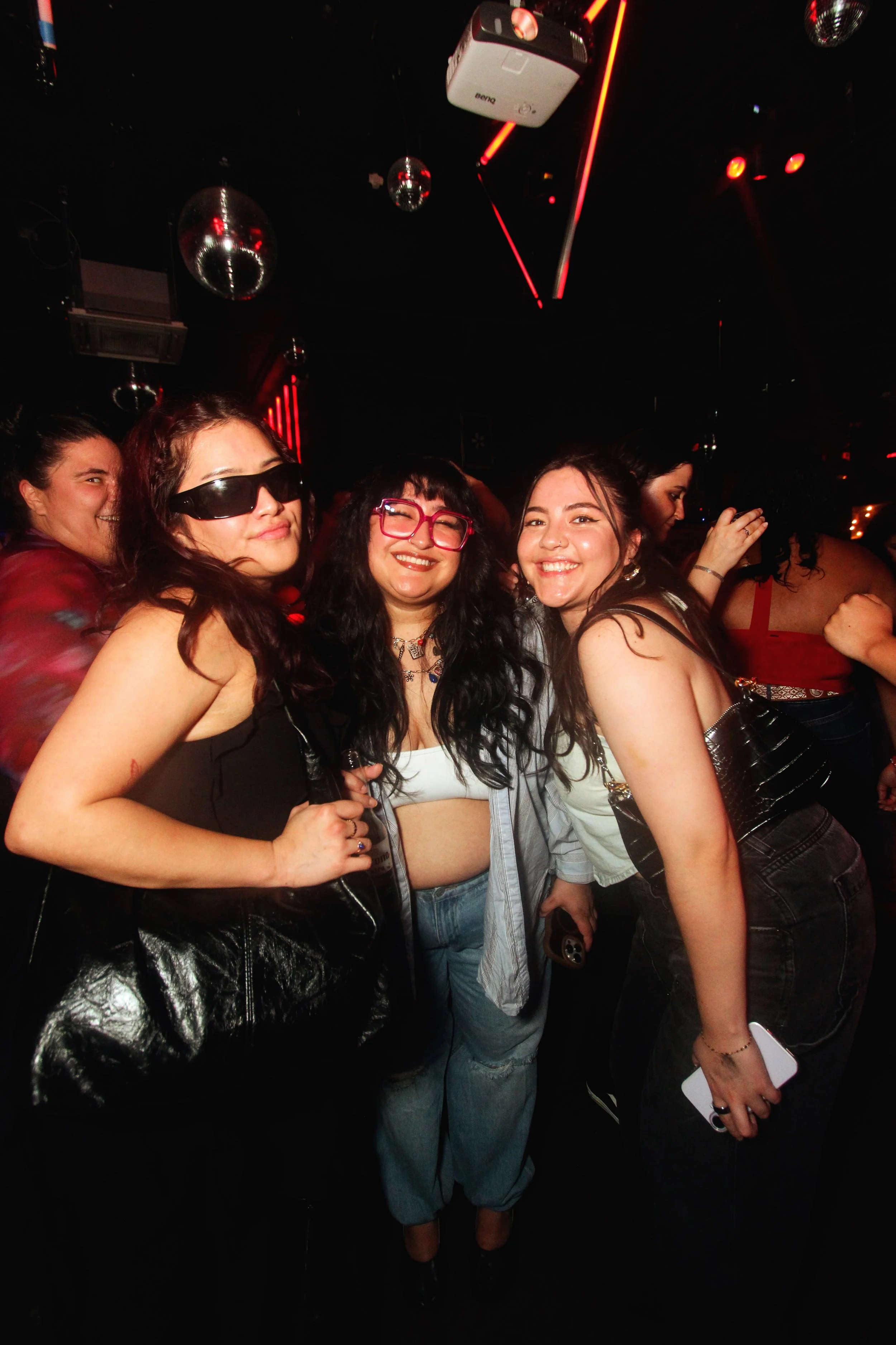 Group of three women smiling and posing at a nightclub with disco balls and red neon lights.