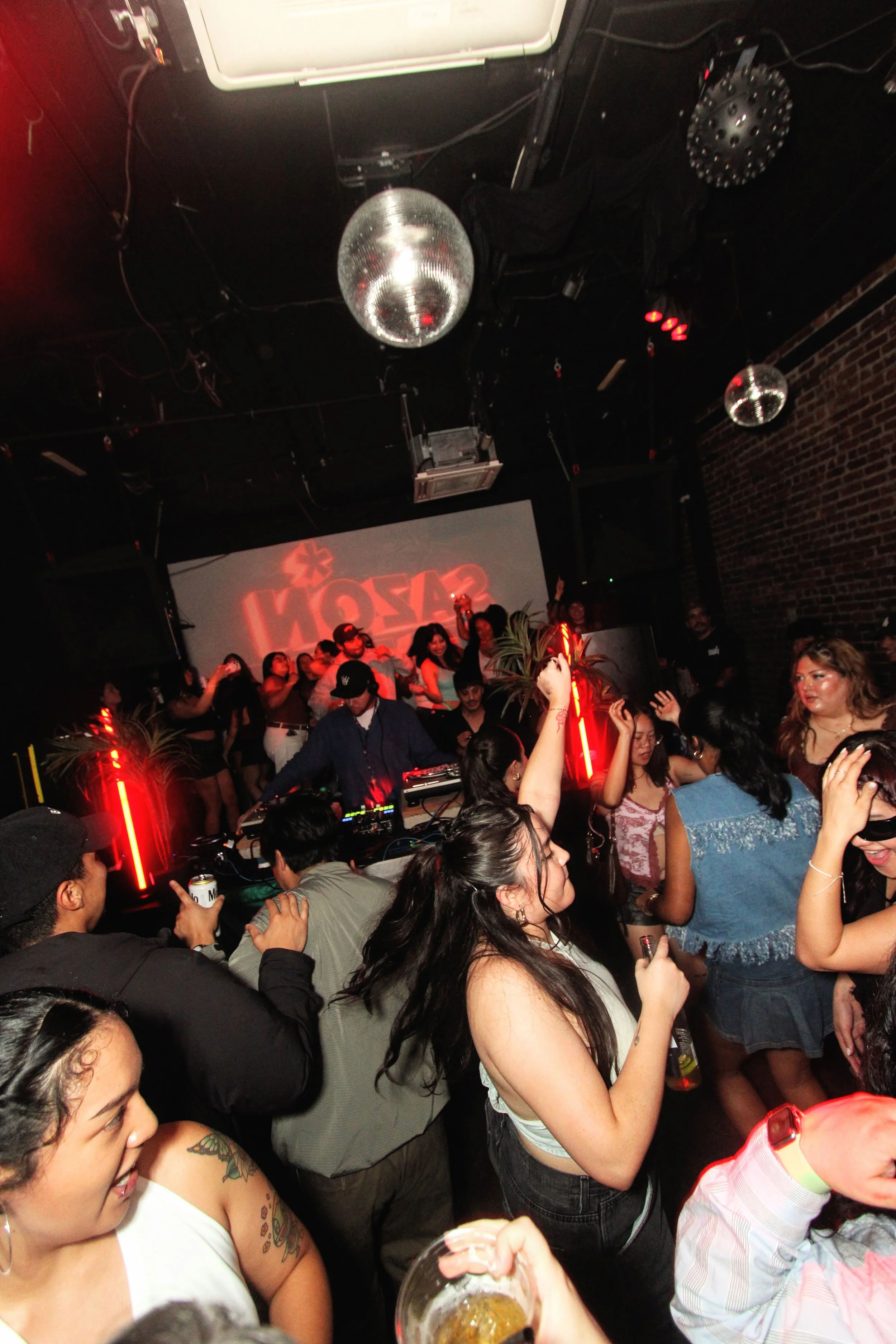 People dancing and socializing at a lively nightclub with a DJ on stage, red neon lighting, and disco balls reflecting light.