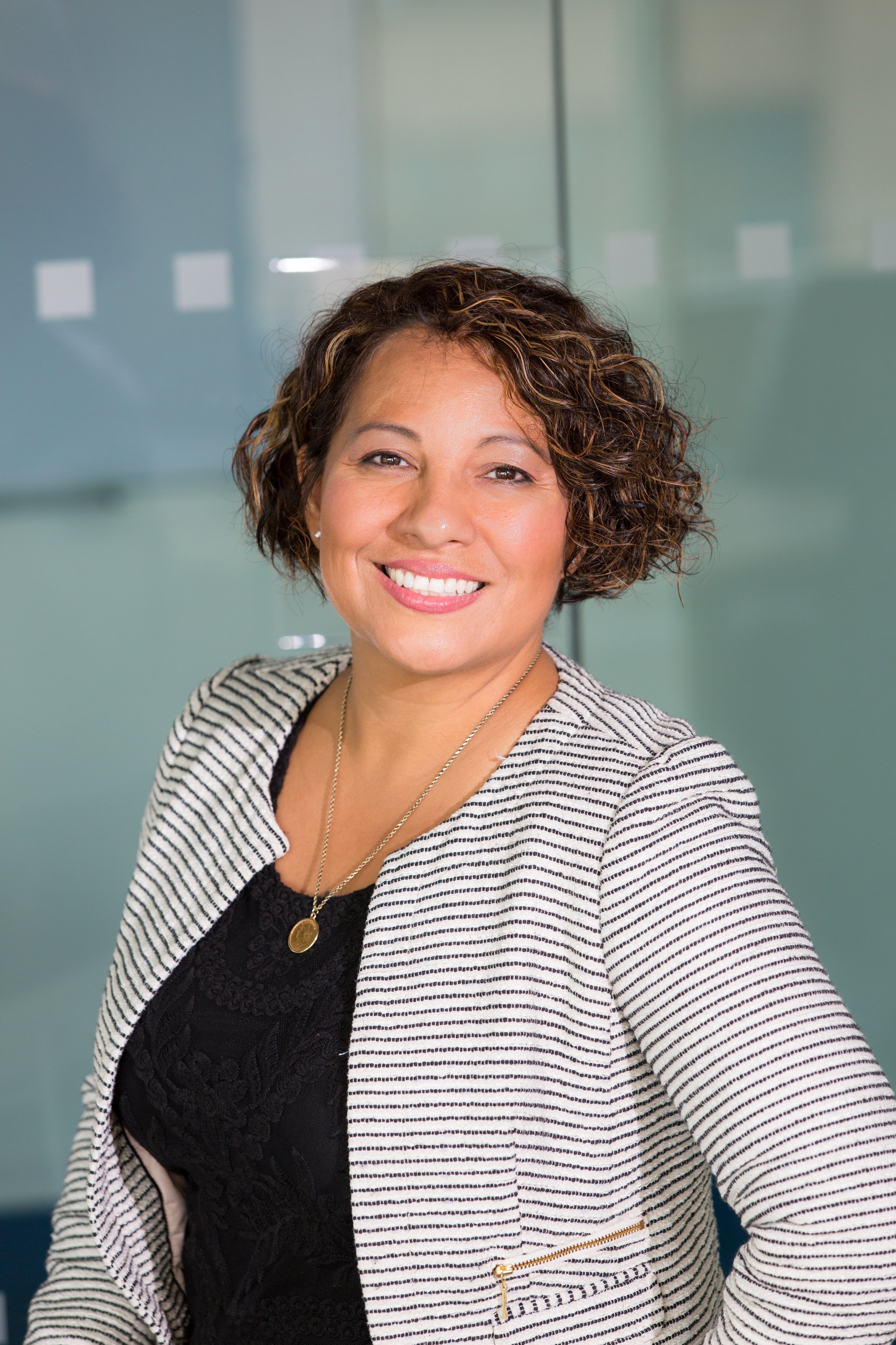 A professional woman with short curly hair, smiling, wearing a black top and striped blazer, standing in an office setting.