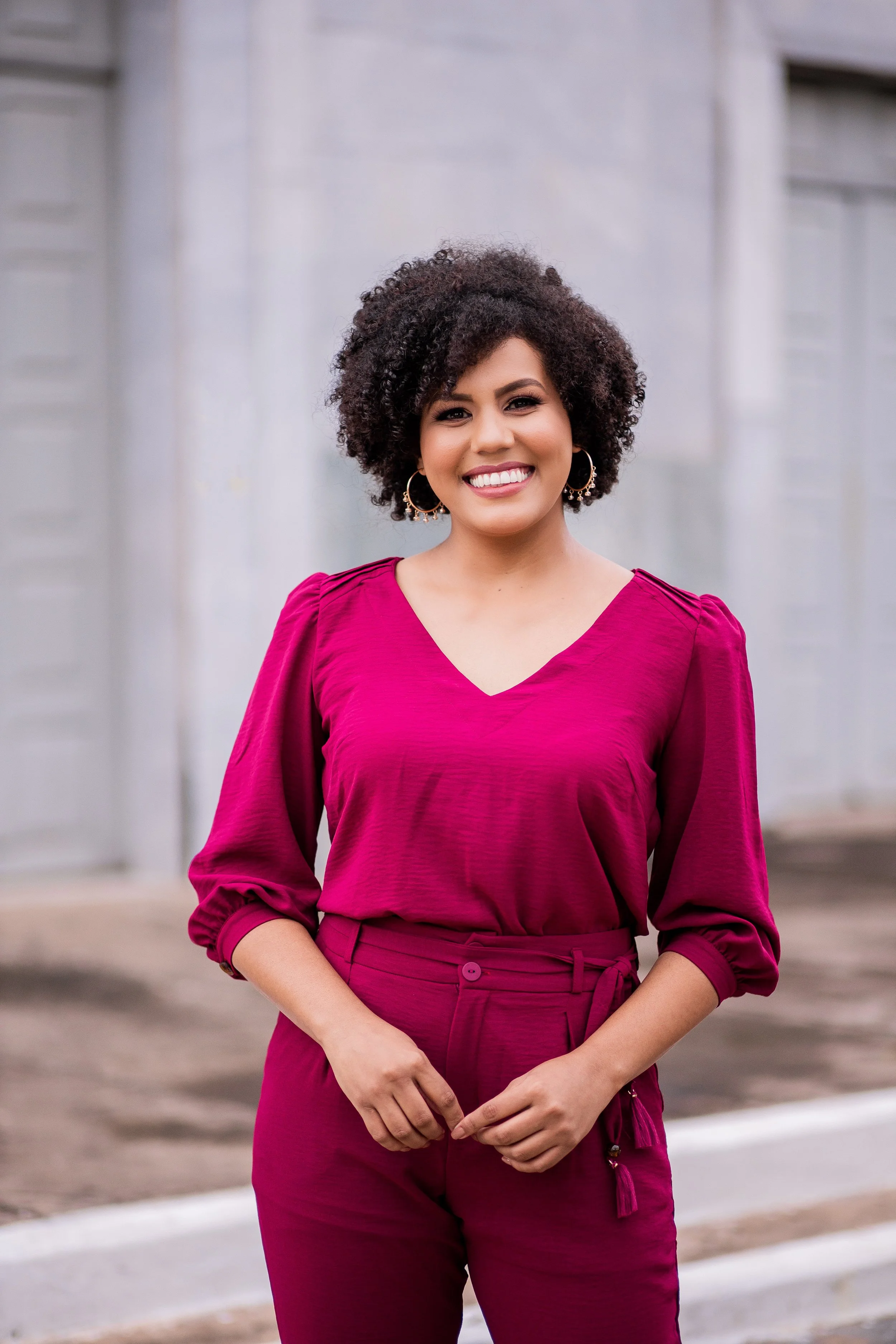 A woman with curly dark hair smiling outdoors, wearing a magenta long-sleeve blouse and matching high-waisted pants, standing in front of a light-colored building.