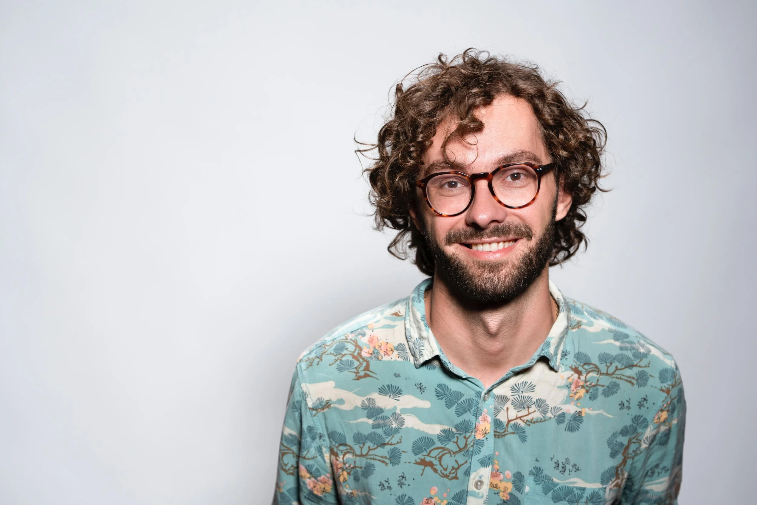 A smiling man with curly brown hair, glasses, and a beard, wearing a blue patterned shirt, standing against a plain light gray background.