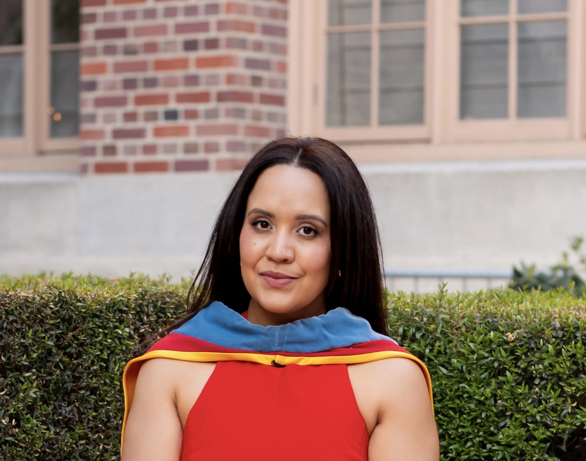 Young woman in a red dress and graduation gown sitting outside with a brick building in the background.