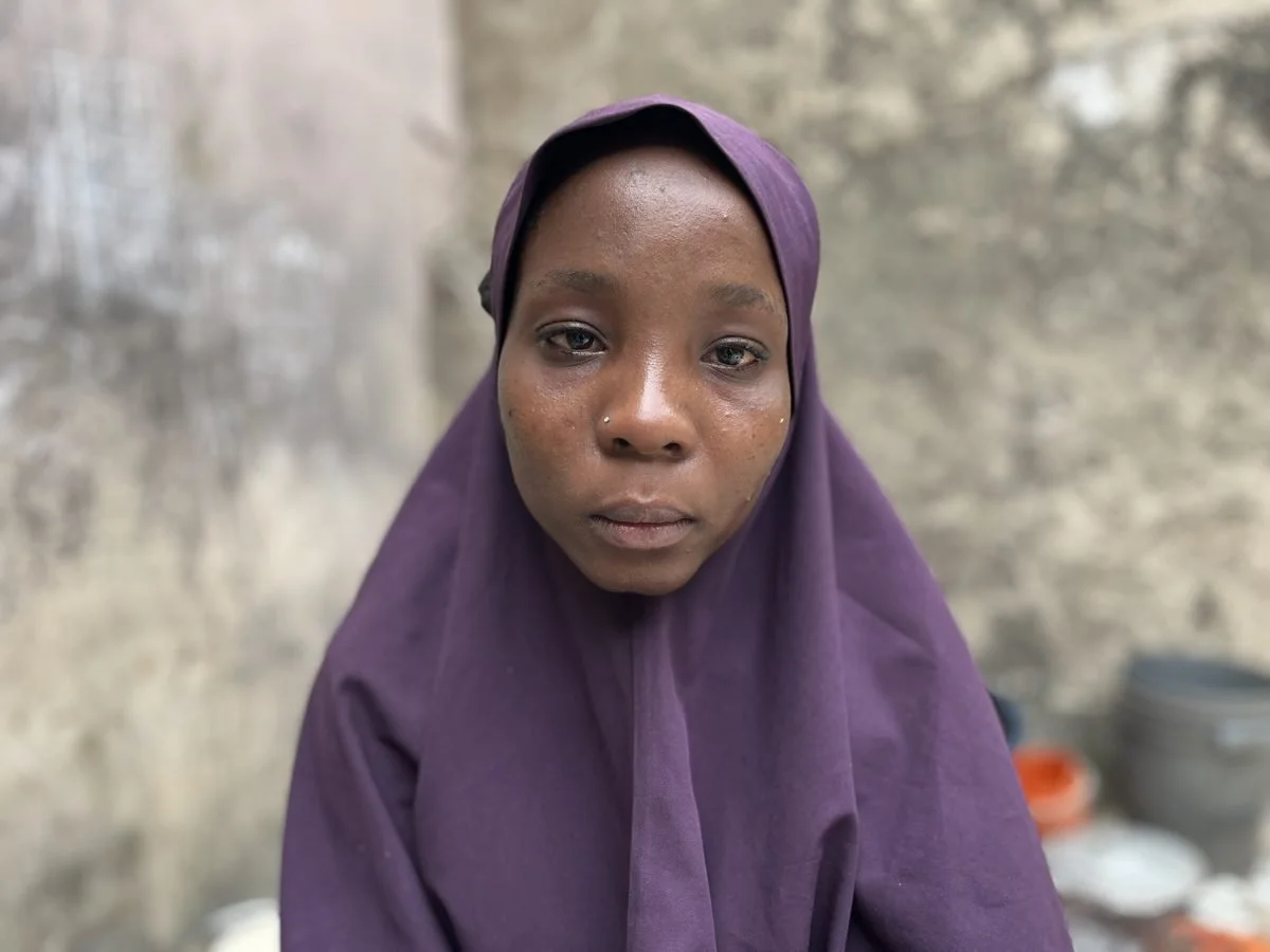 A woman with dark skin wearing a purple headscarf and a nose piercing, sitting in front of a blurred stone wall background.