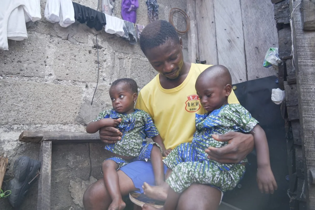 A man holding two young children in a rustic, outdoor setting with laundry hanging above and a wooden wall behind them.