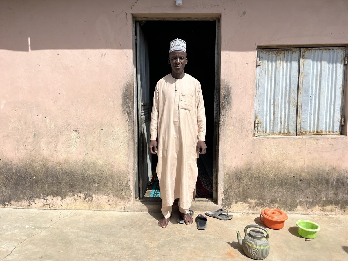 A man in traditional dress stands at the doorway of a house with a pink wall, with various household items on the ground, including a teapot, a green bowl, an orange container, a shoe, and a black shoe. The man is smiling slightly and wears a white cap.