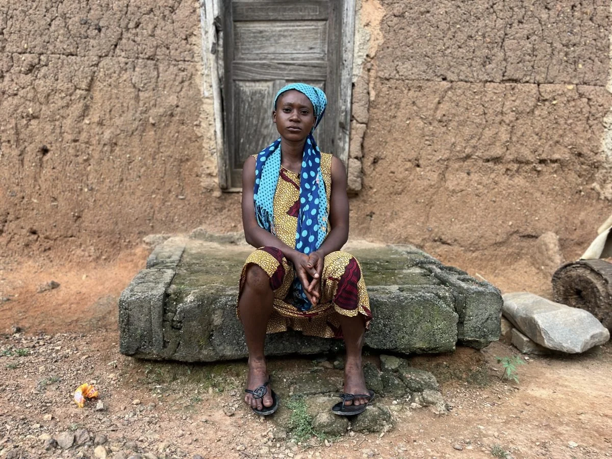 A young girl sitting on a stone step in front of a rustic wooden door with a mud-brick wall background. She is wearing a colorful dress and a headscarf, with her hands clasped in her lap, looking directly at the camera.