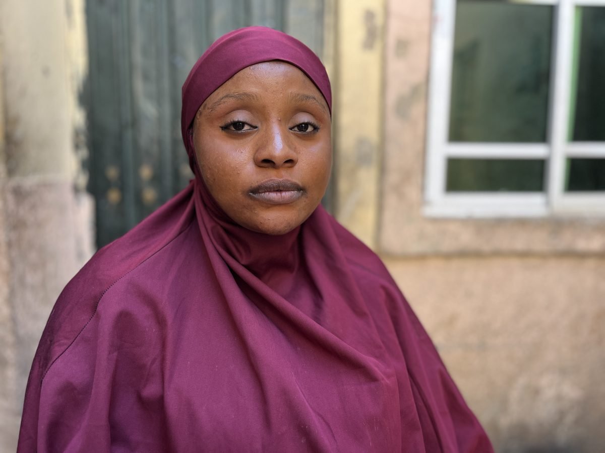 A woman wearing a maroon headscarf and matching clothing standing outdoors in front of a brick and window background.
