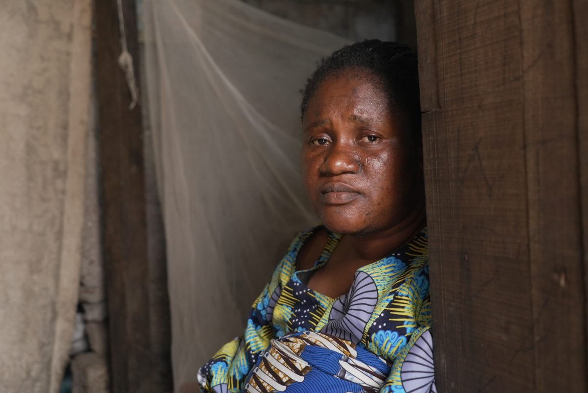 Woman with short curly hair looking out from a narrow space, wearing a colorful patterned dress, inside a wooden and concrete structure.