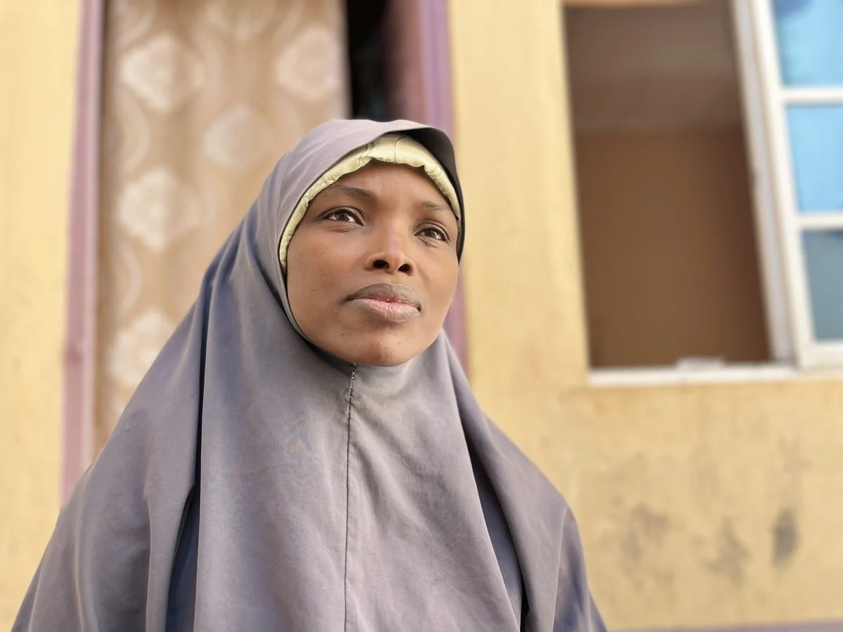 A woman wearing a grey hijab standing inside a room with yellow walls and a window.