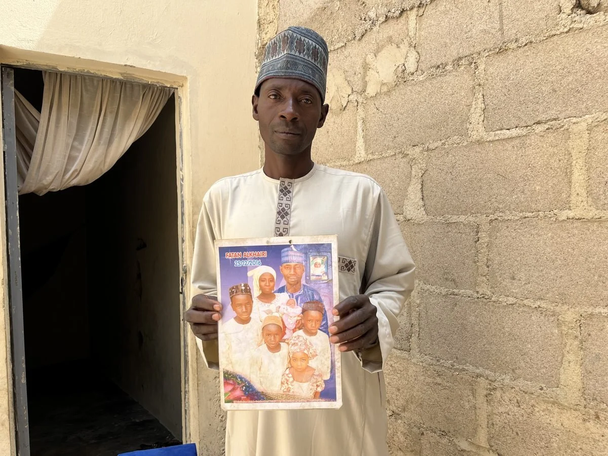 A man dressed in traditional attire holding a family portrait in front of a stone wall, with a doorway and curtain behind him.