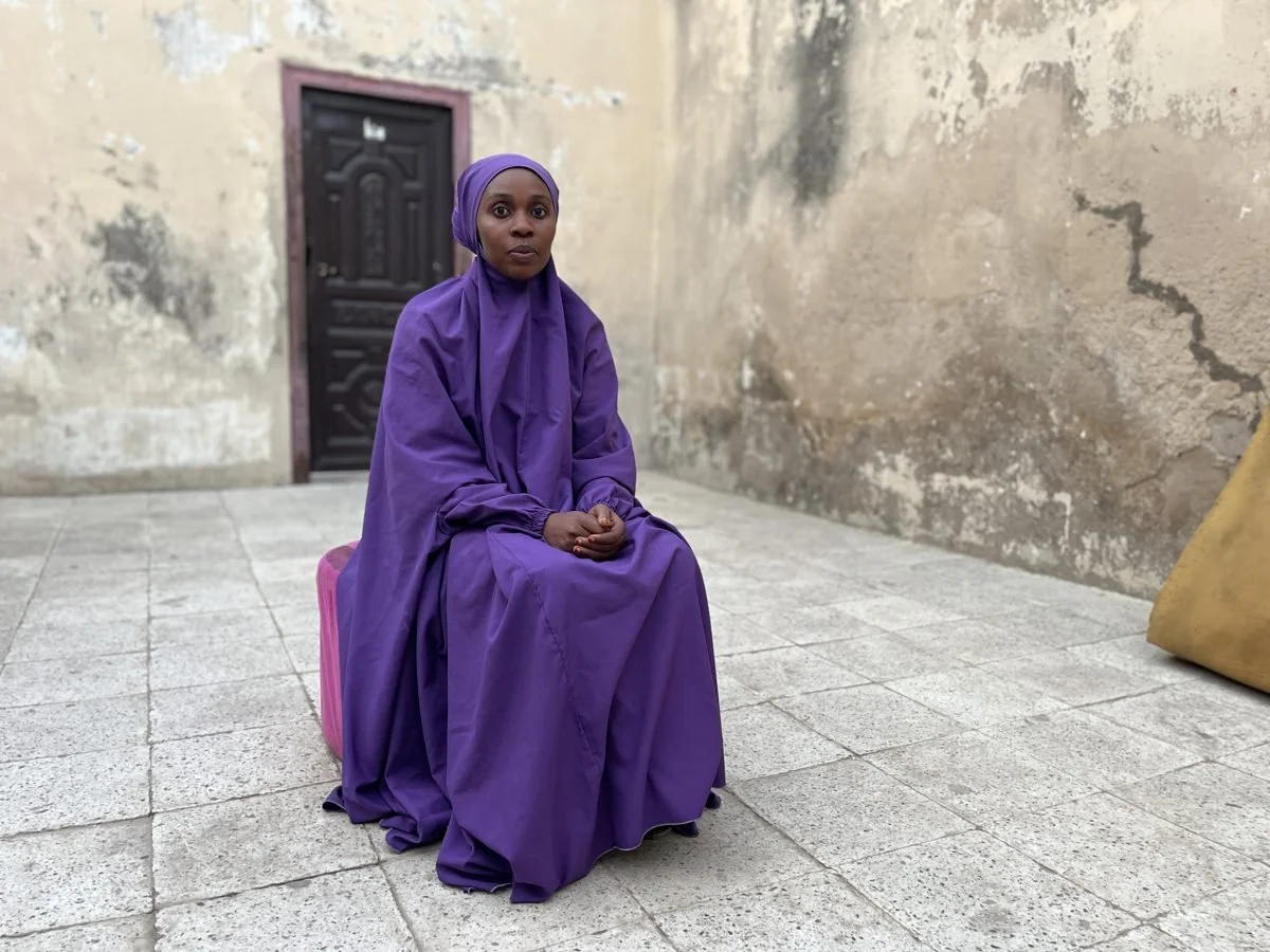 A woman in a purple dress and headscarf sitting on a pink suitcase outside near a weathered beige wall with a dark door.