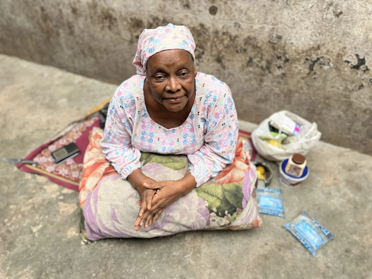 Elderly woman sitting outdoors on a sack with her hands clasped, surrounded by personal belongings including a plastic bag, a jar, and packaged food items.
