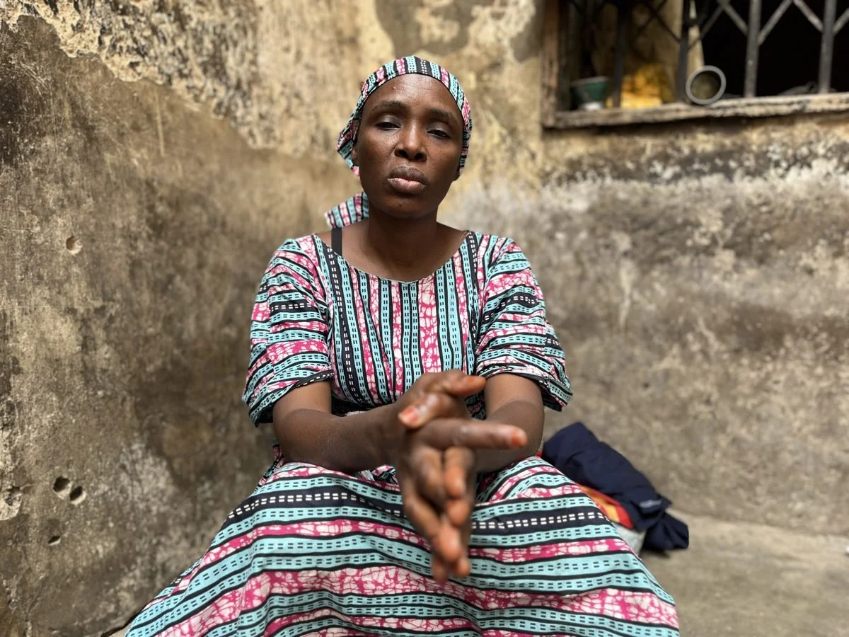 Woman in colorful patterned dress and headscarf sitting against a weathered wall with her hands clasped, outdoors.