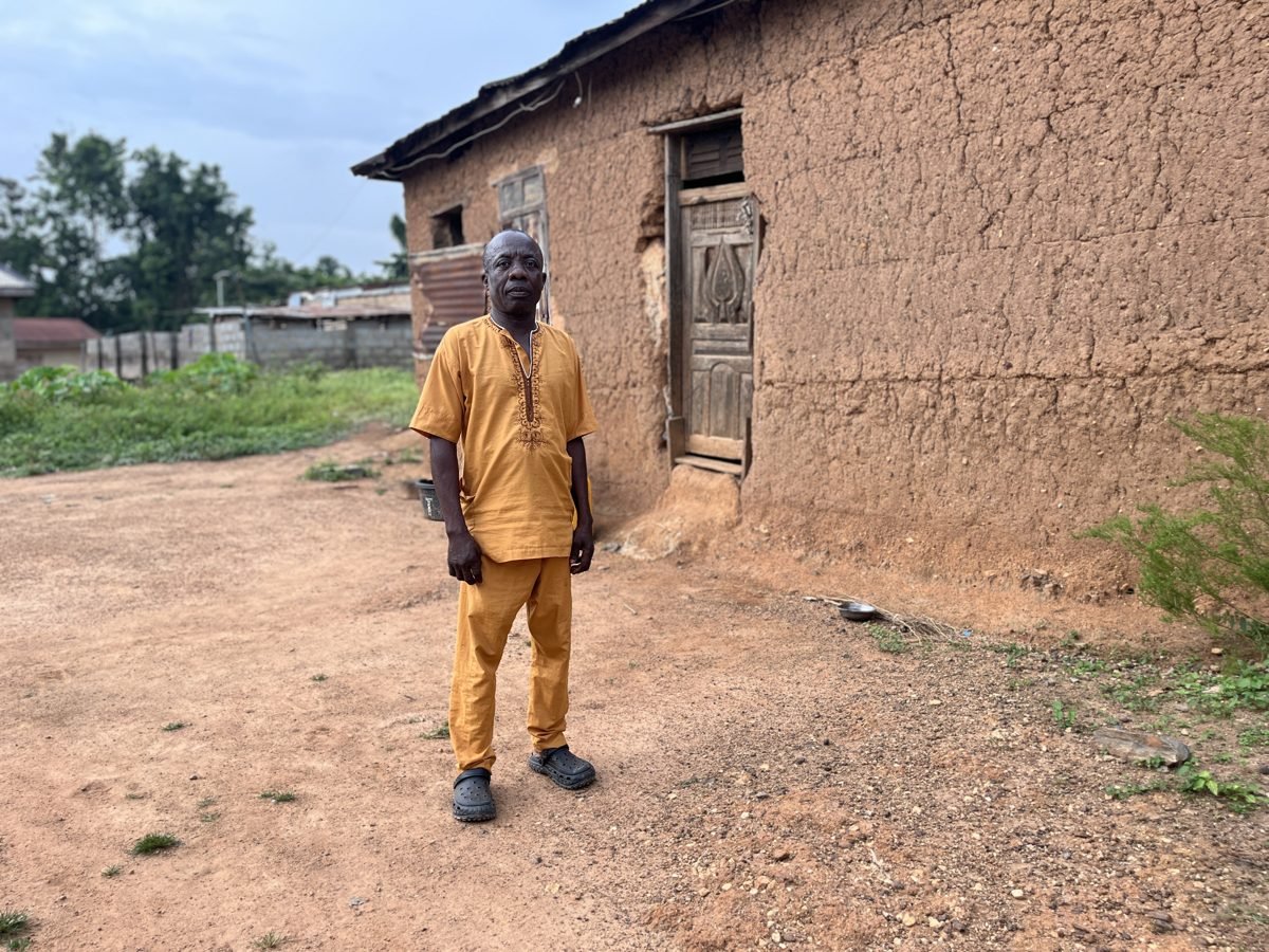 A man in a yellow traditional outfit standing outside in front of a mud brick house with a wooden door.
