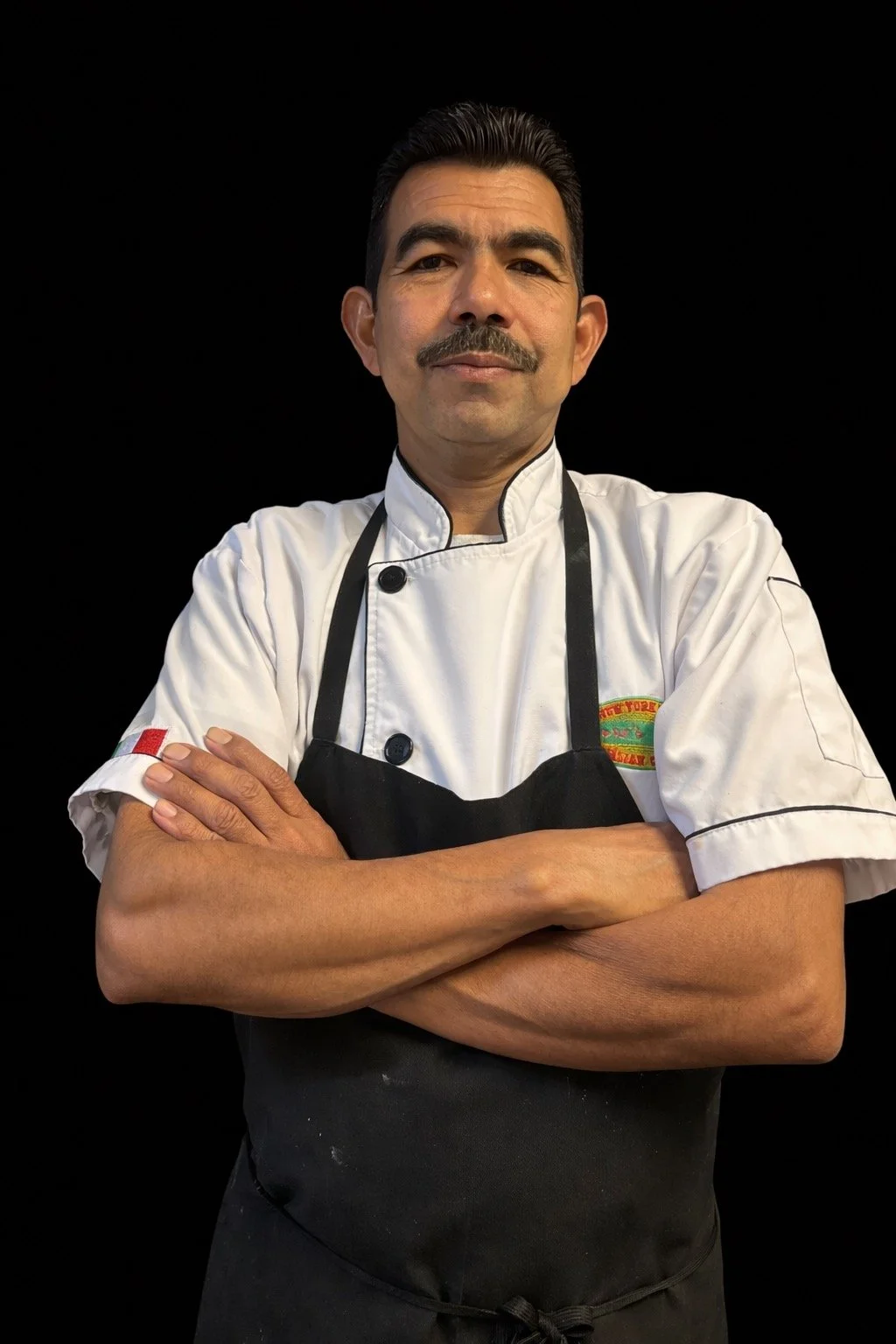 A male chef with a mustache, wearing a white chef's coat with a colorful patch on the sleeve, a black apron, and a wireless microphone headset, standing with arms crossed against a black background.