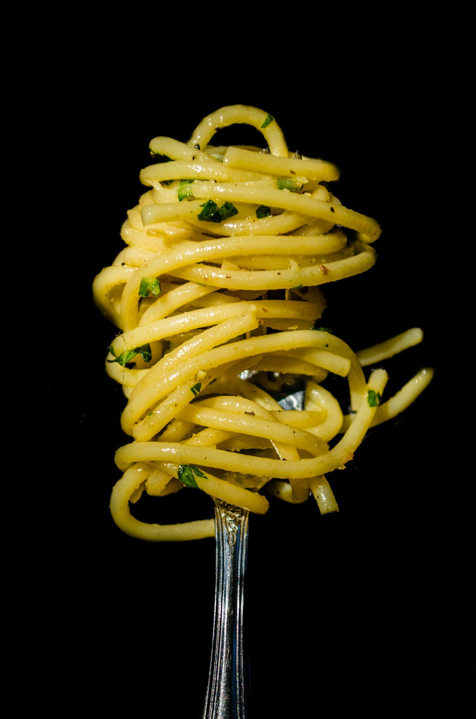 Spaghetti noodles garnished with chopped herbs on a silver fork against a black background.