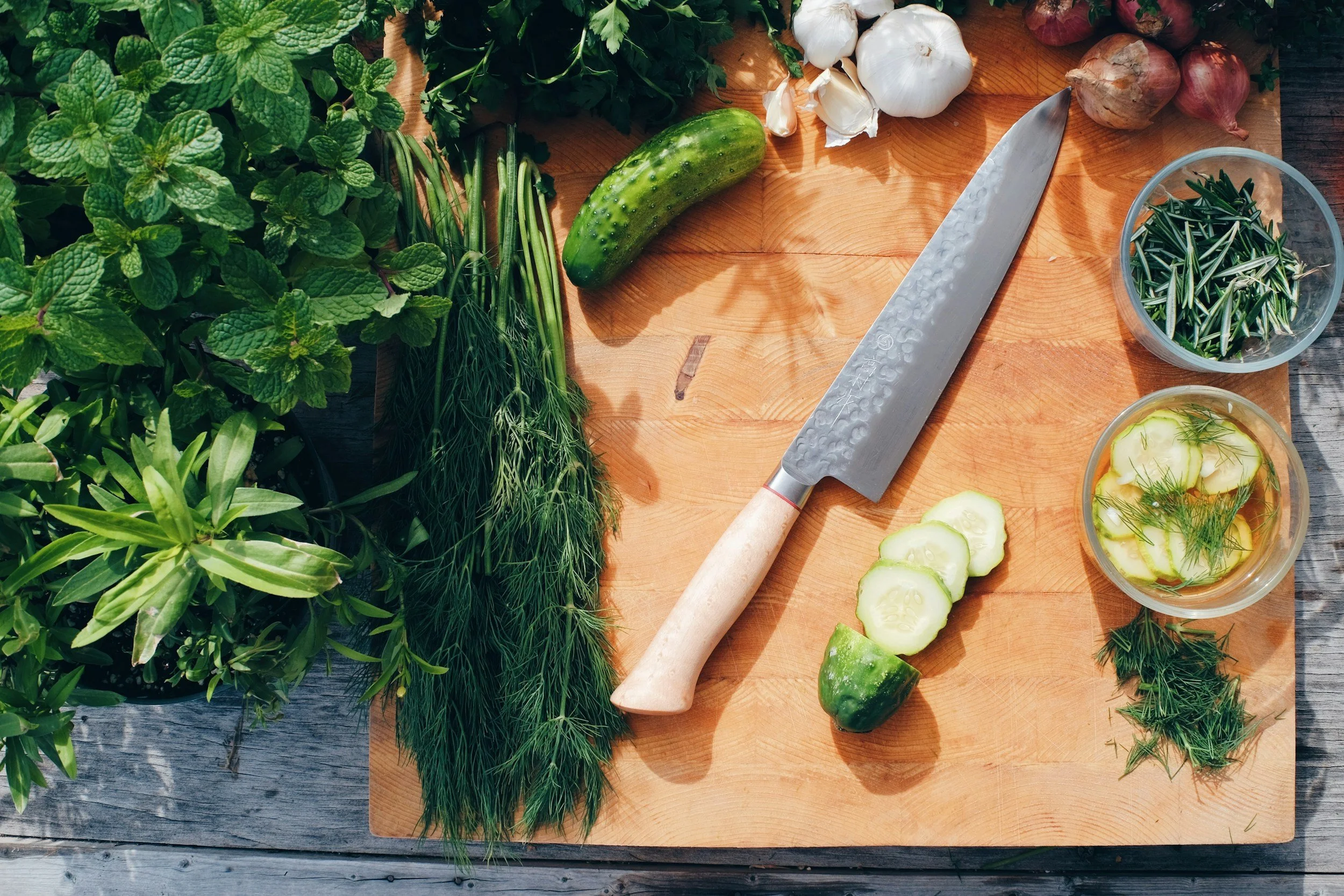 Fresh herbs, vegetables, and a knife on a wooden cutting board, including mint, dill, cucumber, garlic, shallots, and a bowl of cucumber slices in vinegar with dill, outside on a wooden surface.