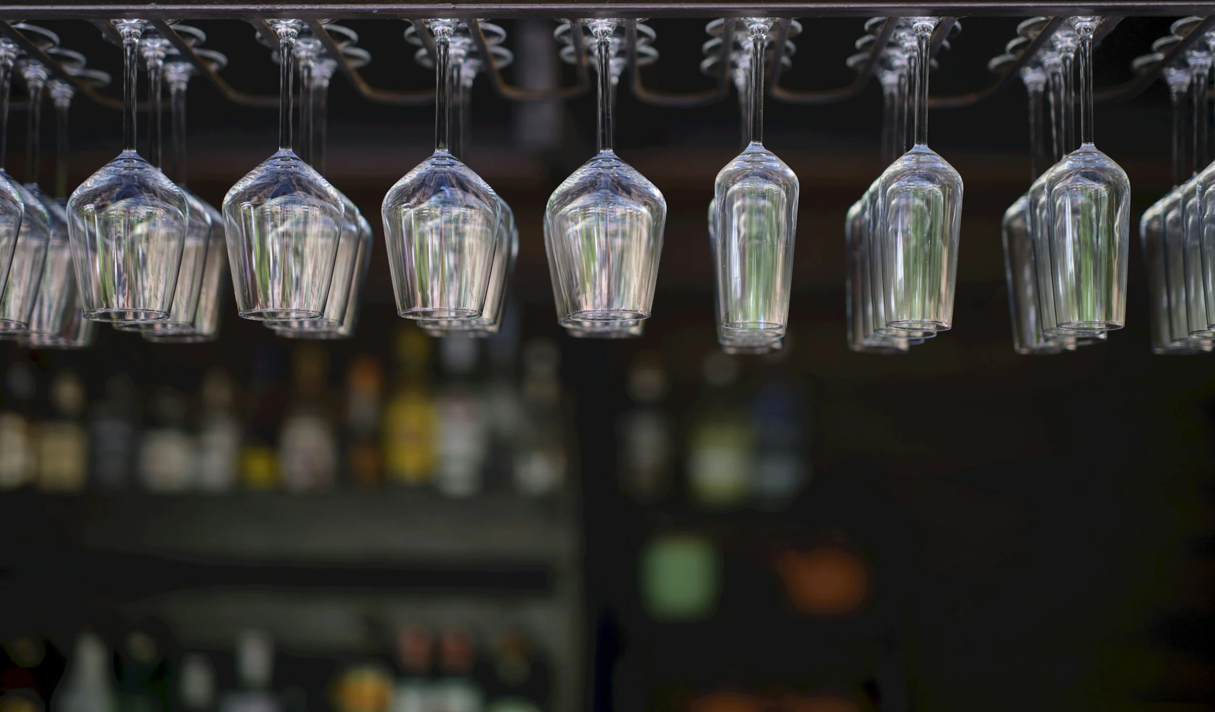 Empty wine glasses hanging upside down above a bar counter with bottles in the background.