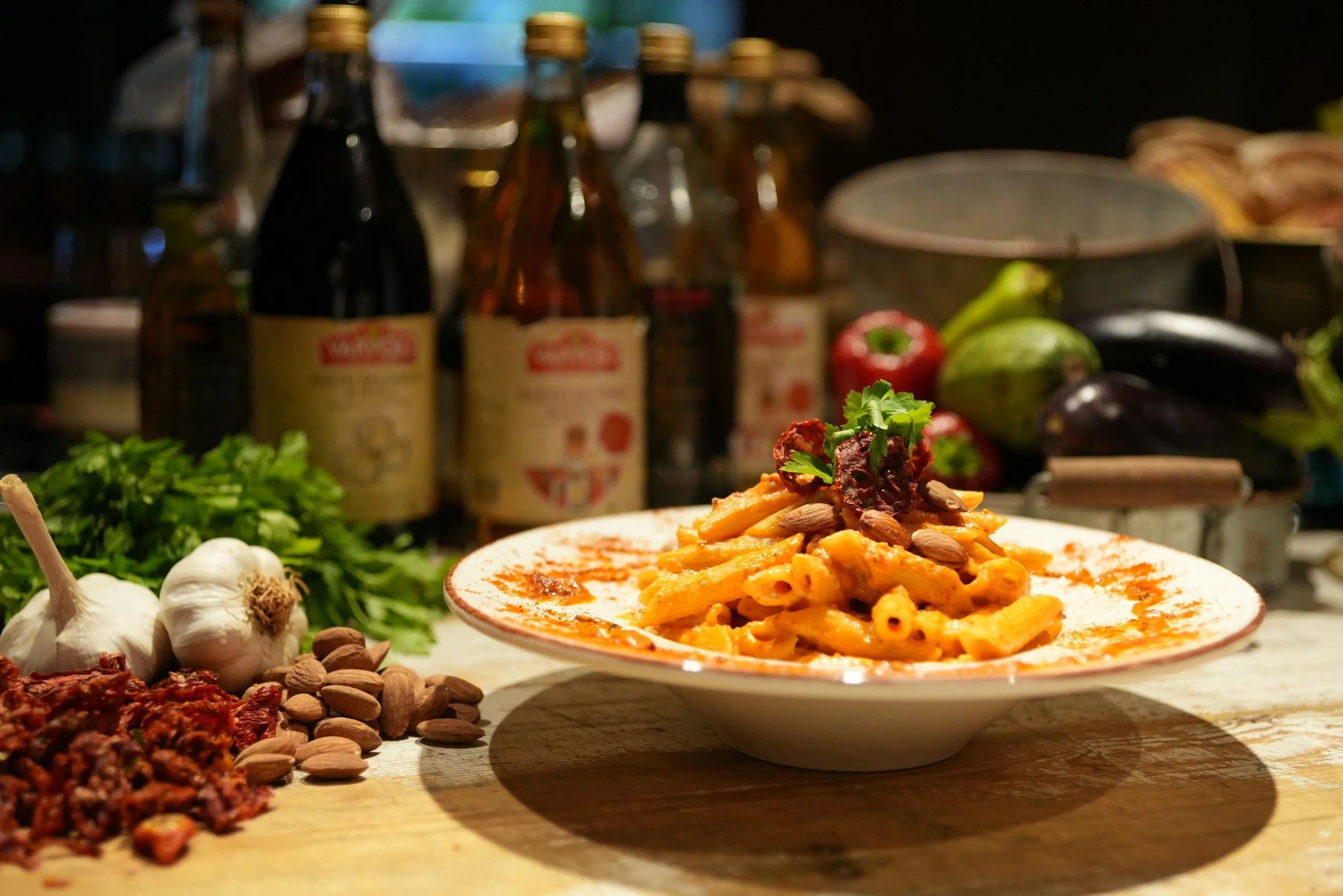Plate of pasta with tomato sauce garnished with herbs and almonds, surrounded by garlic, basil, almonds, and ingredients for cooking on a kitchen countertop, with bottles and vegetables in the background.