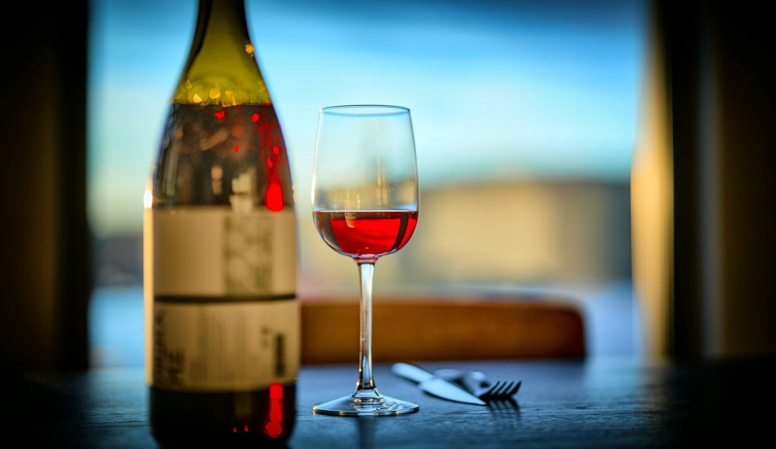 A wine glass filled with red wine, a wine bottle, a fork, and a knife on a table against a blurred background with a window and bright sky.